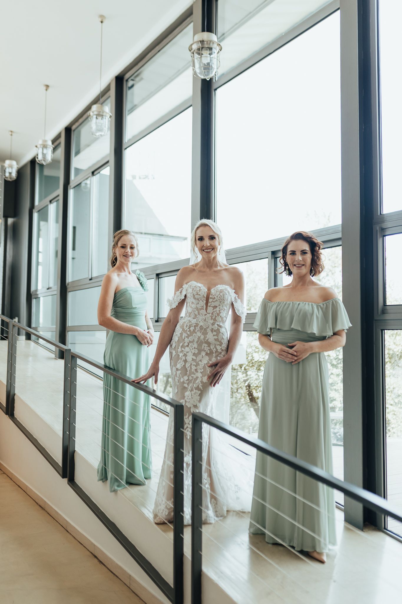 Bride and bridesmaids in mint green dresses standing by large windows in modern venue.