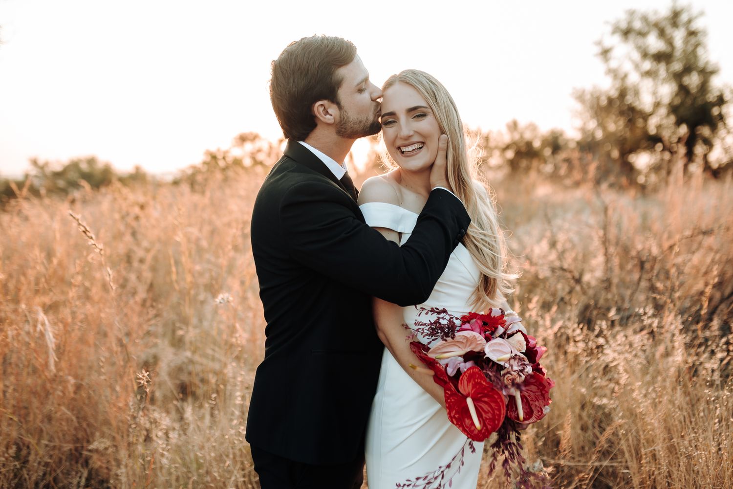 Wedding couple sharing tender moment in autumn field with red bouquet.