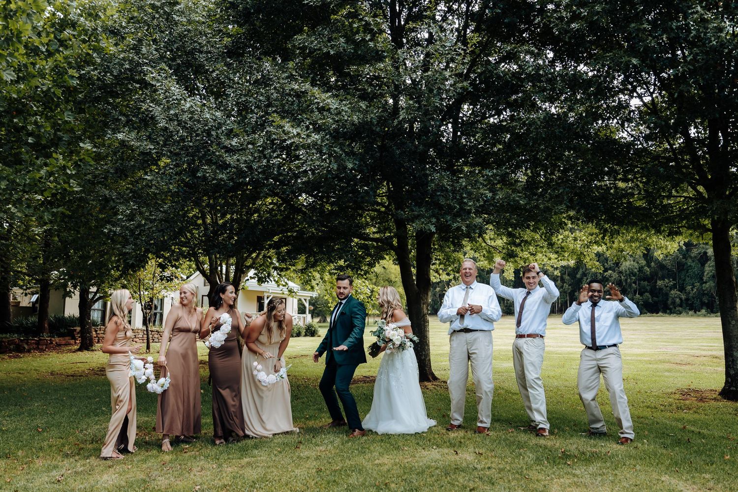 A group of wedding guests walking in a line through a lush green park during an outdoor summer celebration.