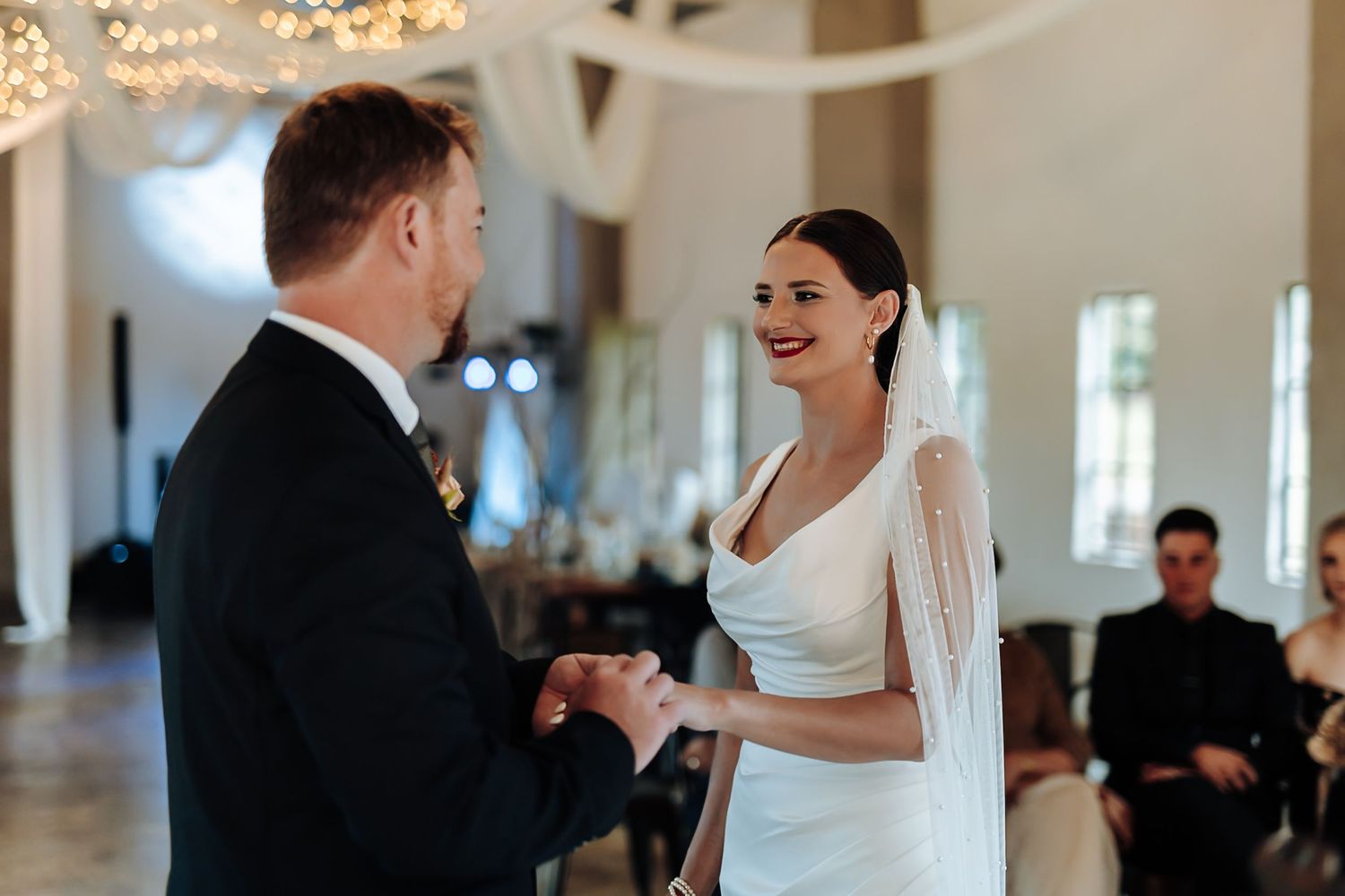 A couple shares their first dance as newlyweds in an elegant ballroom with chandeliers and guests watching.