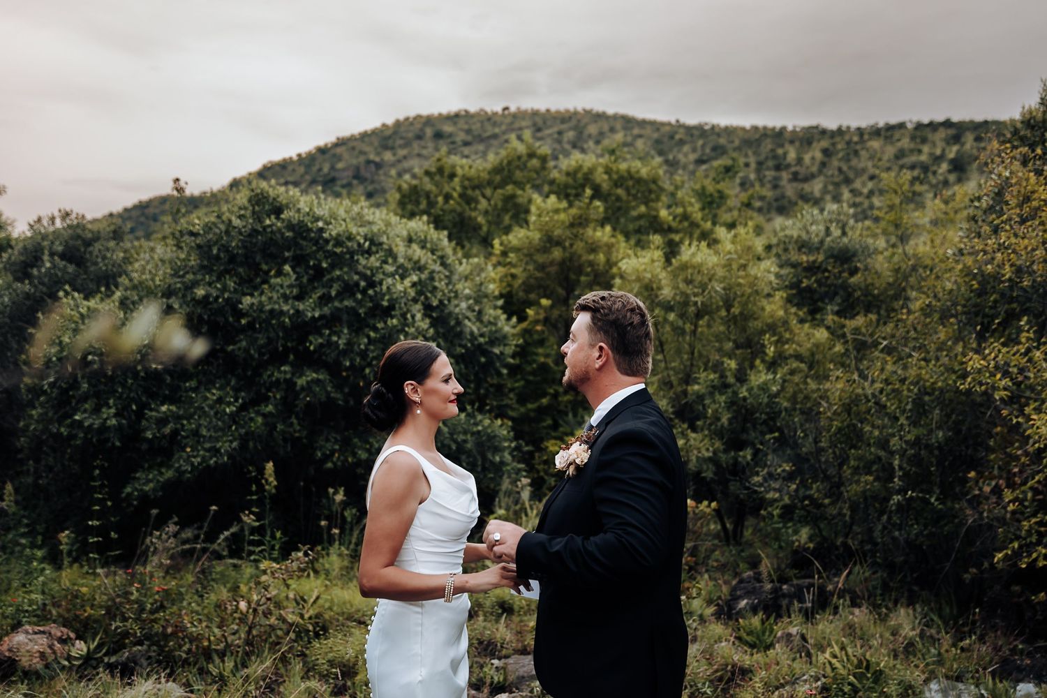 A couple shares a romantic moment during their outdoor wedding ceremony surrounded by lush green mountains and trees.