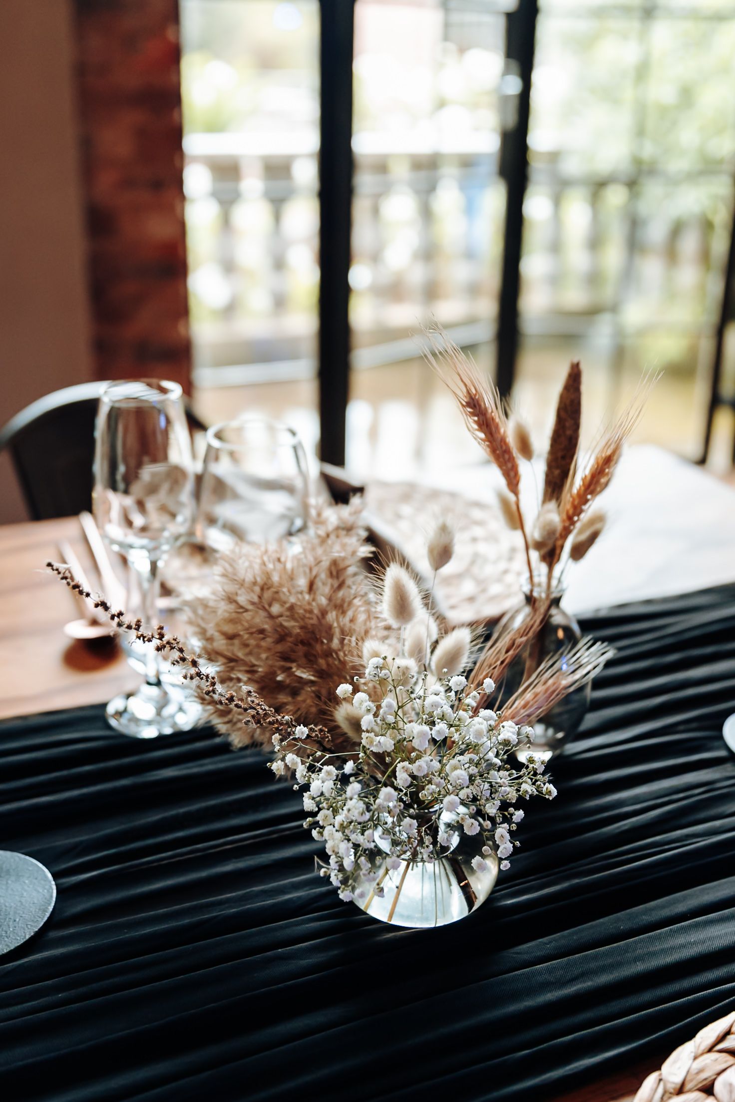 Dried wheat stalks and white baby's breath flowers arranged in clear glass vases on a black tablecloth.