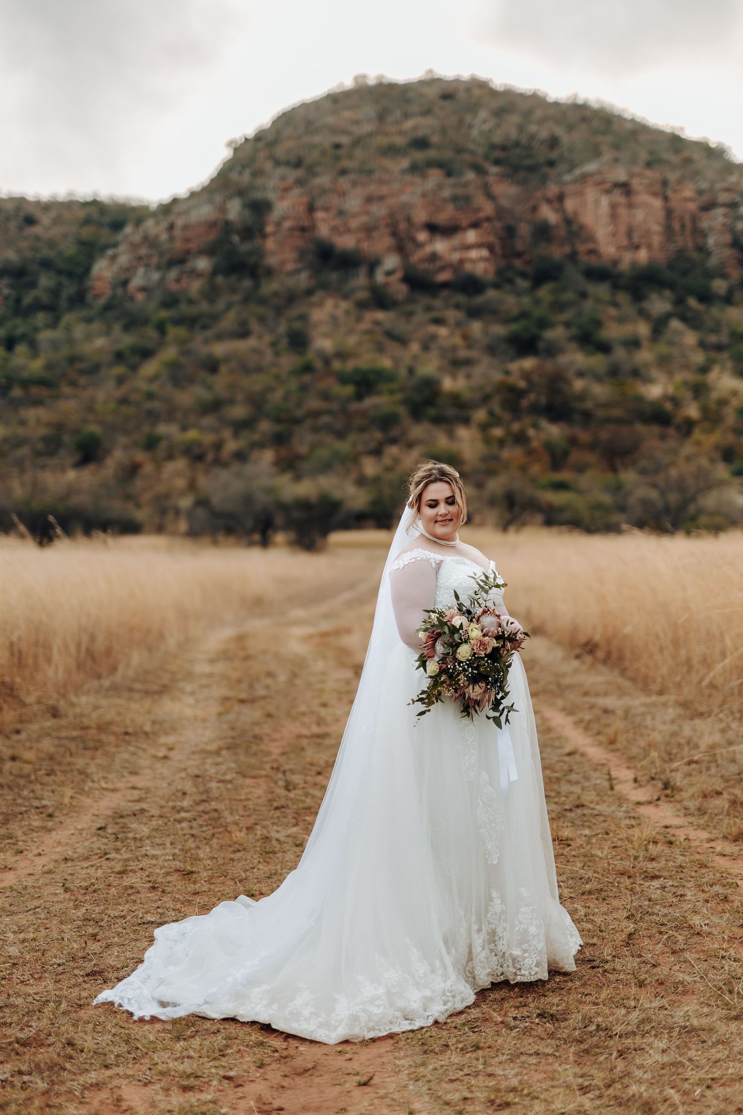 A bride in a flowing white wedding dress holds a bouquet of flowers while standing in a field with mountains in the background.