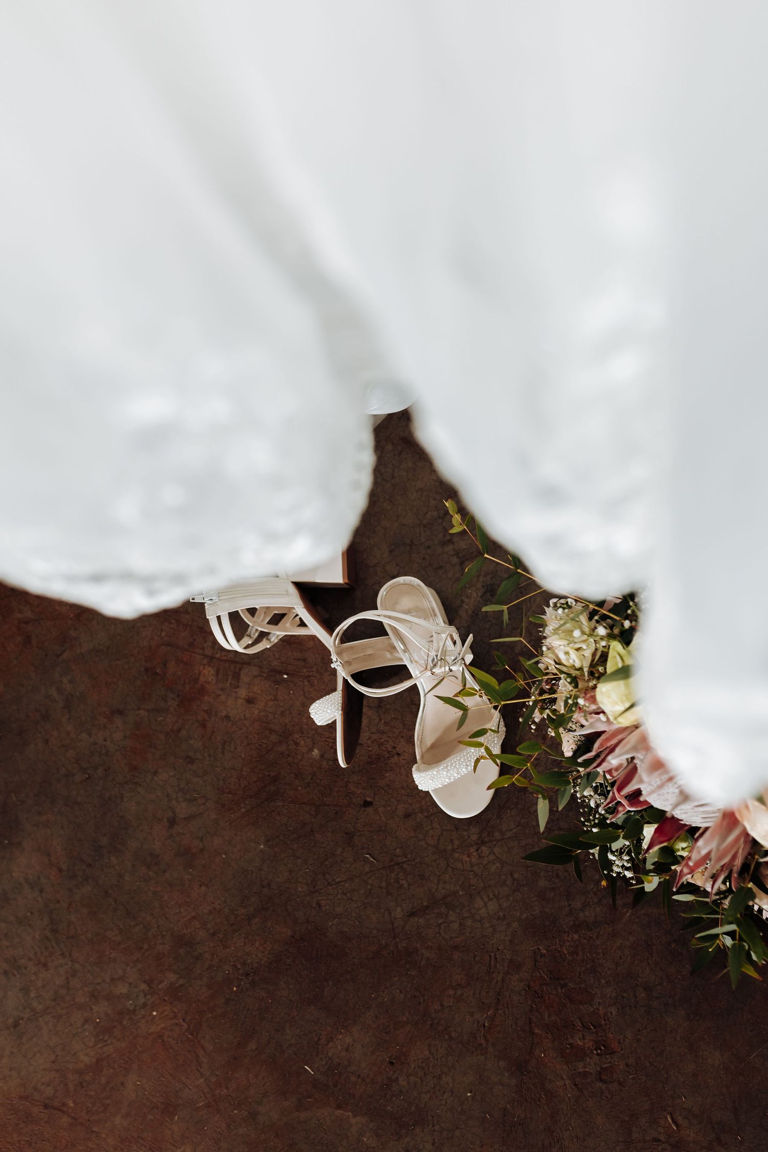 A white bridal high heel shoe and floral bouquet rest on a dark rustic surface against a light background.