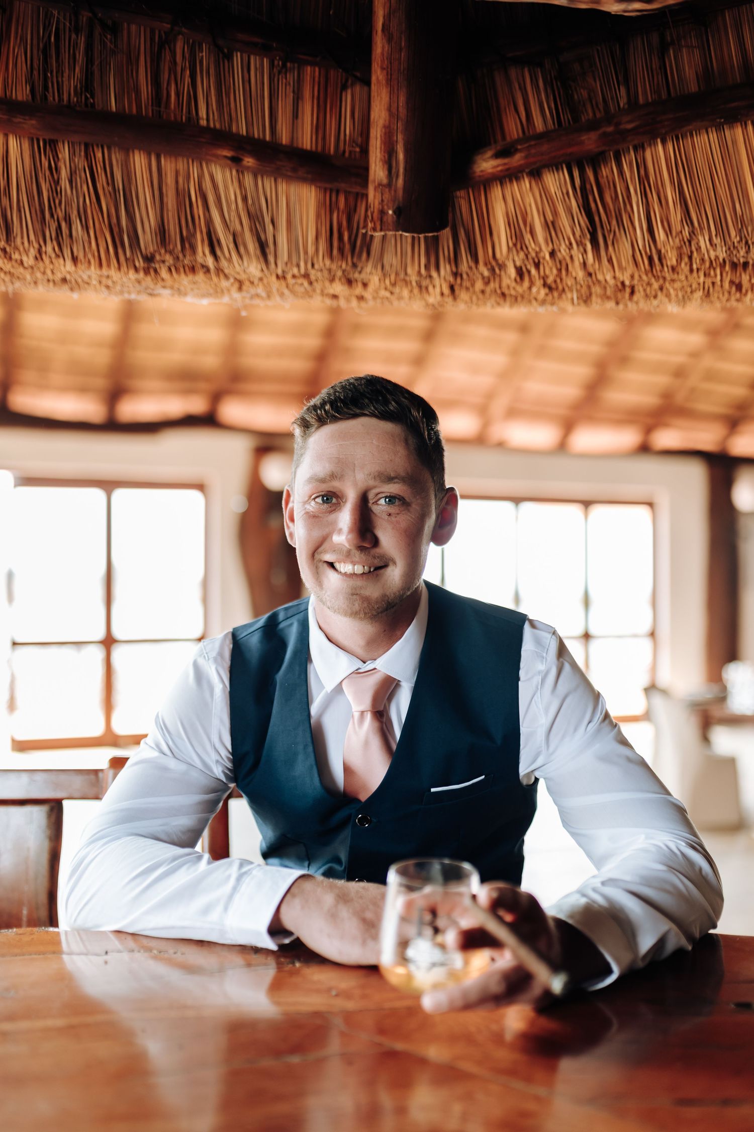 Man in formal attire rests at a wooden bar counter in tropical thatched-roof setting.