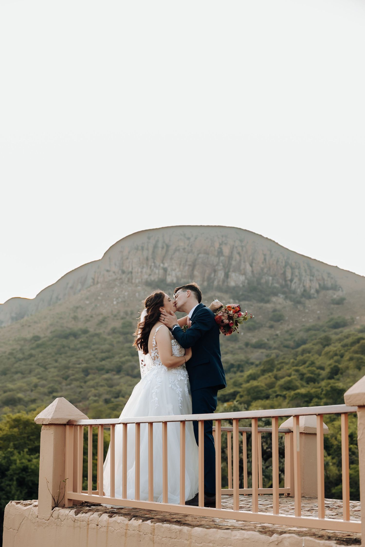 A couple in wedding attire embraces on a wooden deck overlooking scenic mountain landscapes at sunset.