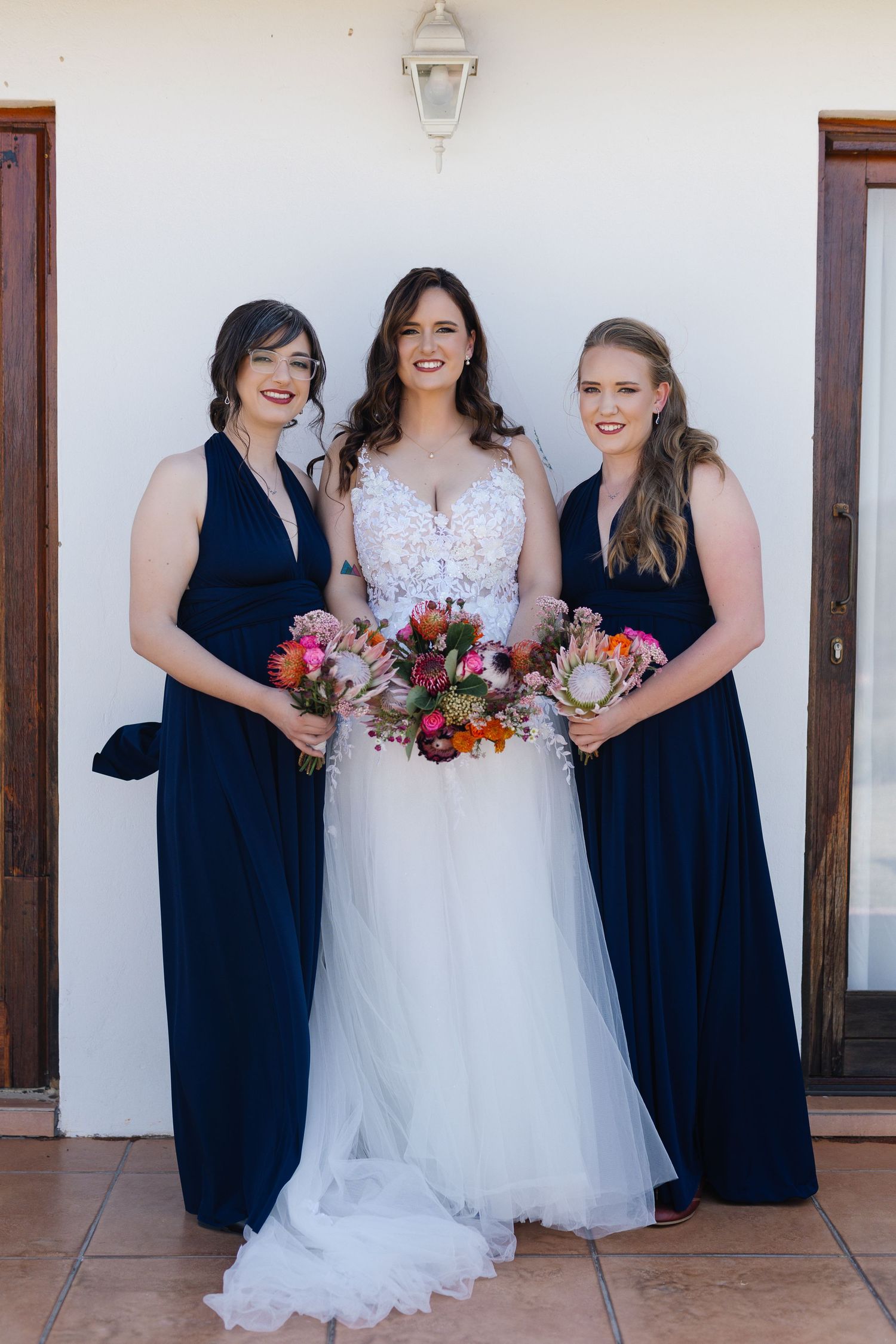 Wedding party wearing navy blue bridesmaid dresses and white bridal gown pose with colorful bouquets against white wall.