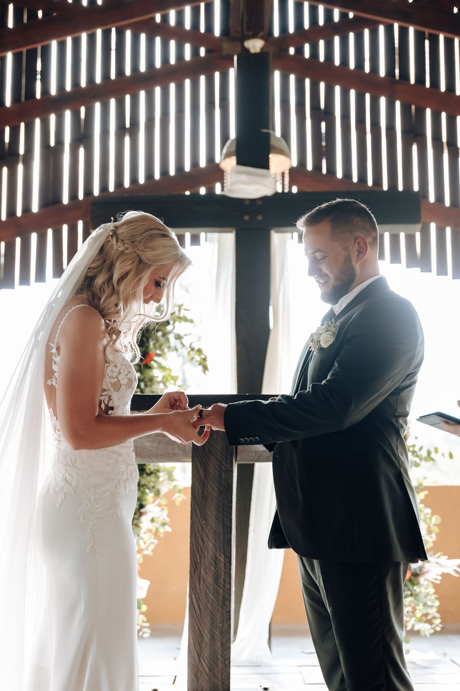 A couple exchanges rings during their wedding ceremony in a rustic wooden barn with sunlight streaming through slats.