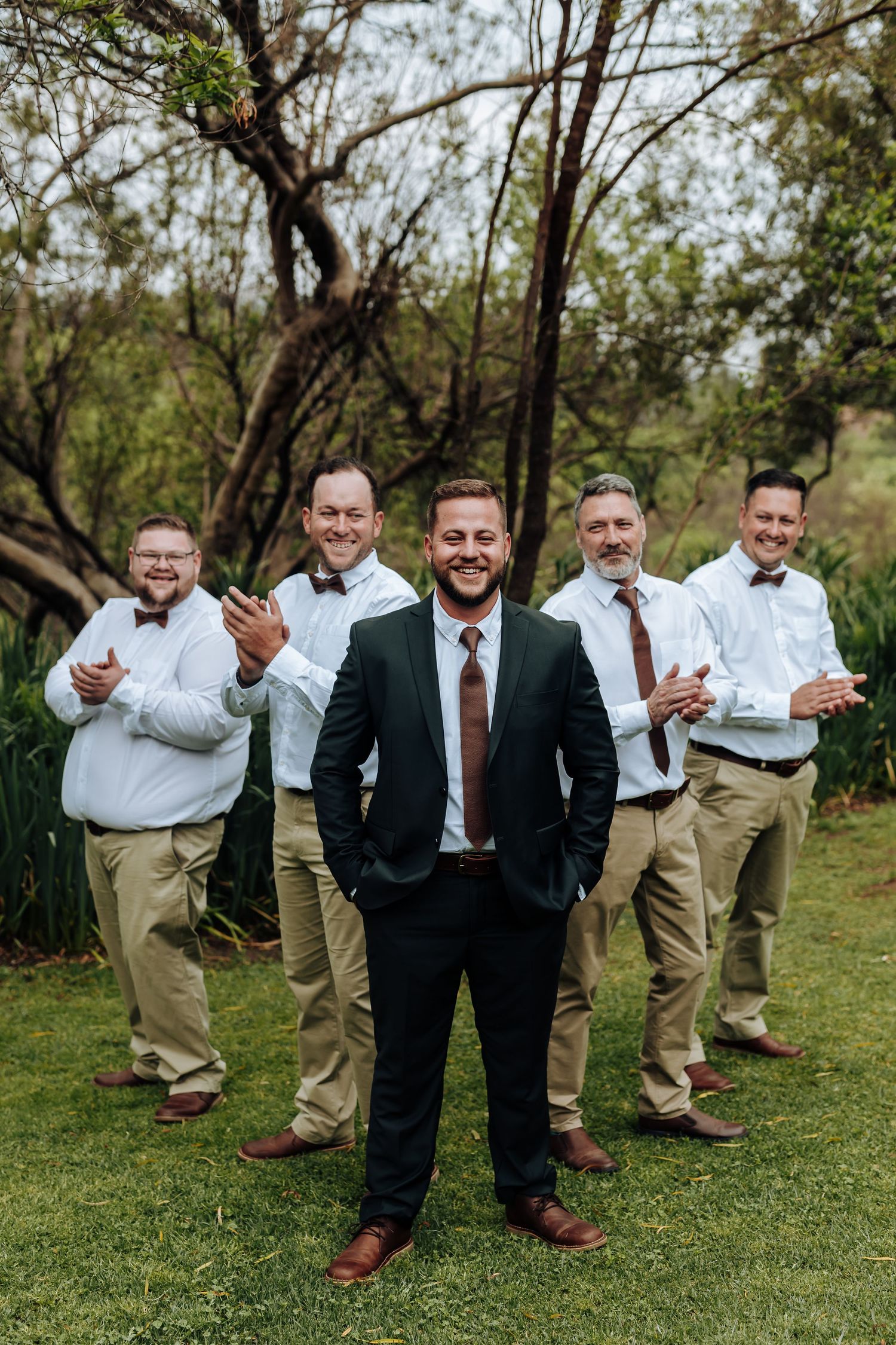 A group in formal attire poses playfully on grass with groomsmen in khakis and dark jackets standing behind suited men.