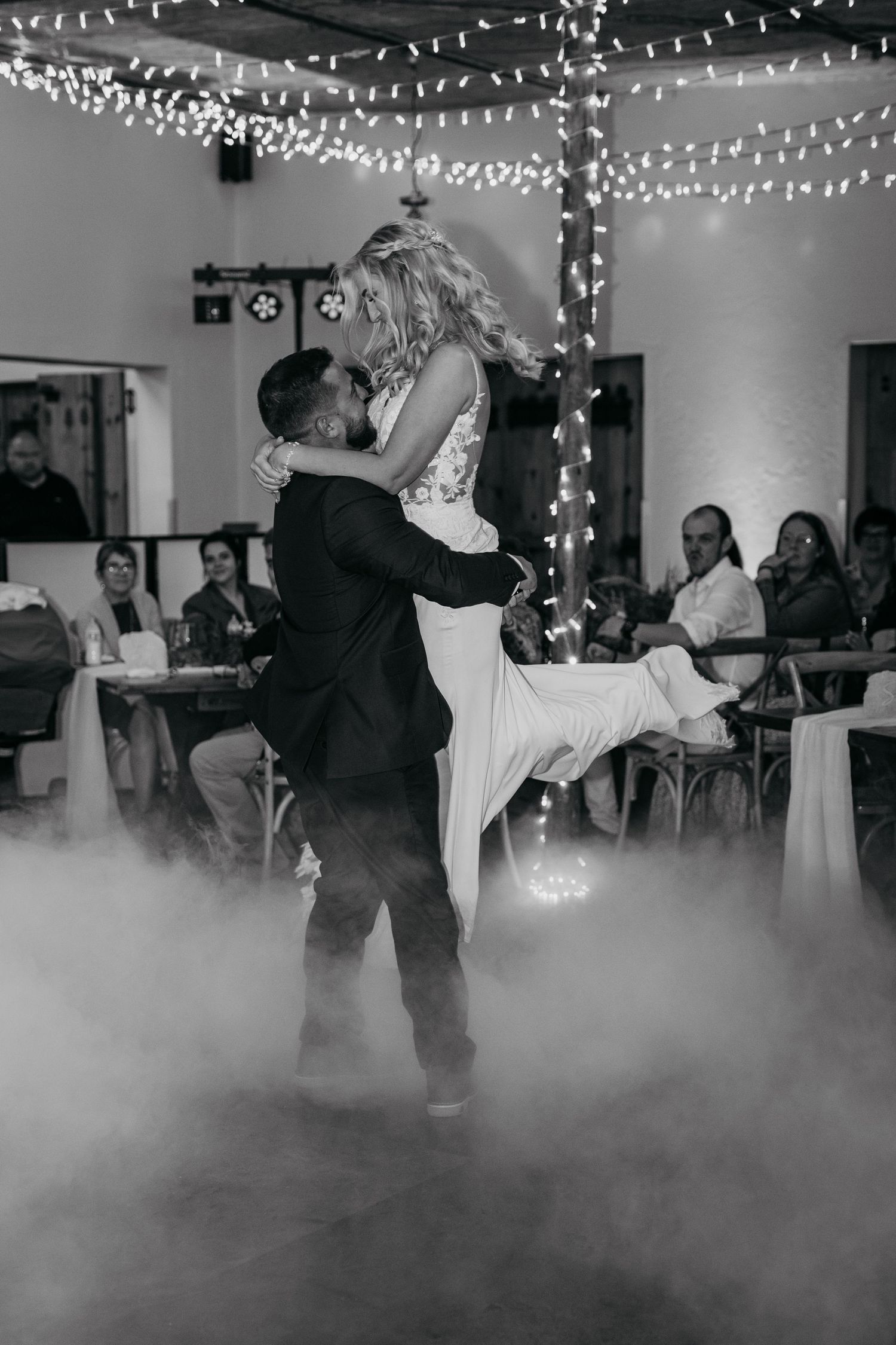 A couple shares their first dance at a wedding reception with fog on the floor and twinkle lights overhead in black and white.