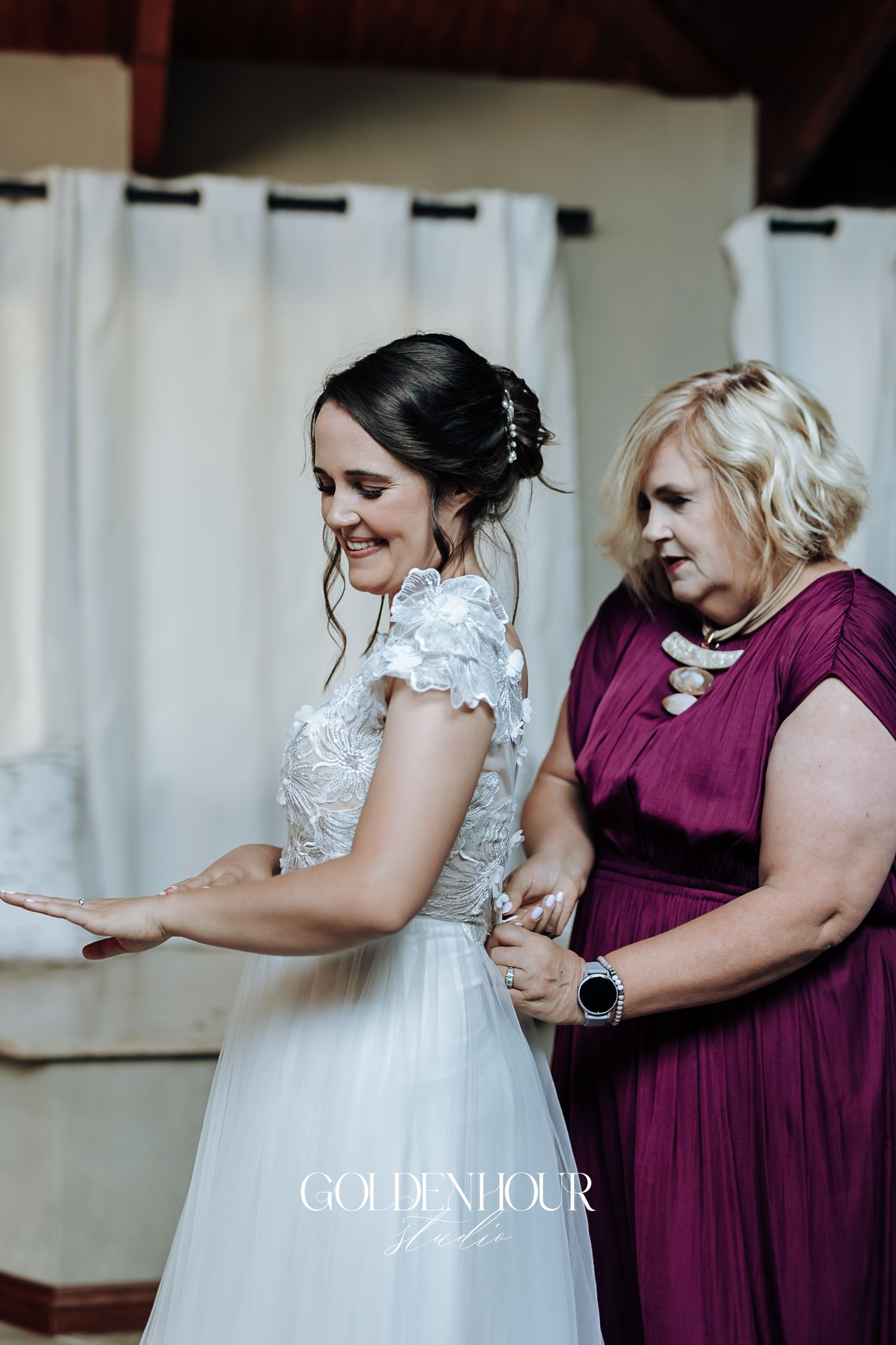 Bride in lace wedding gown being assisted by a woman in a burgundy dress.