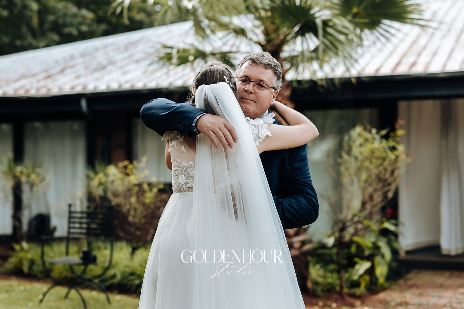 Emotional bride and groom sharing a warm hug outside rustic wedding venue surrounded by greenery.