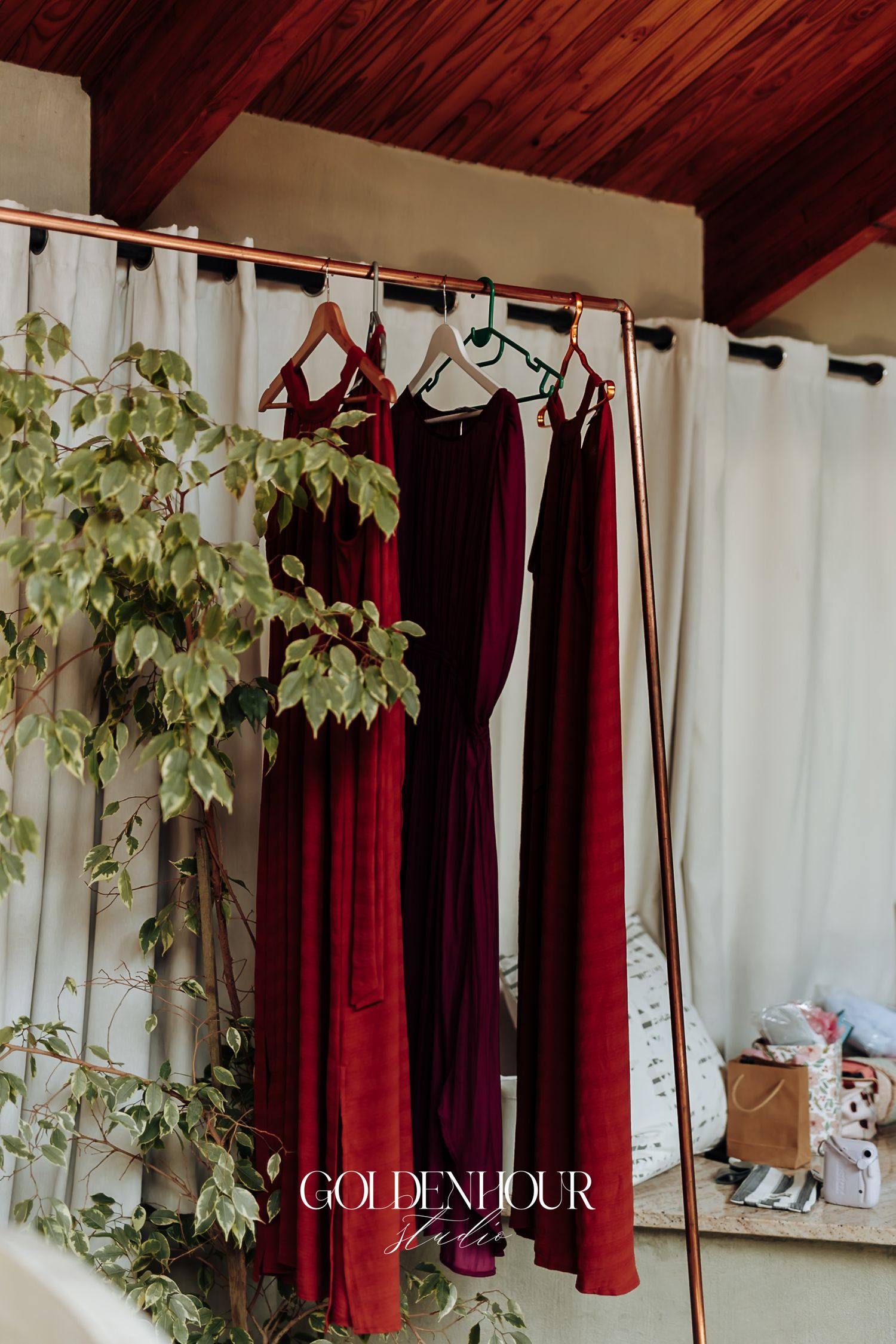 Burgundy and wine-colored bridesmaid dresses hanging on a copper rack beside lush green foliage.