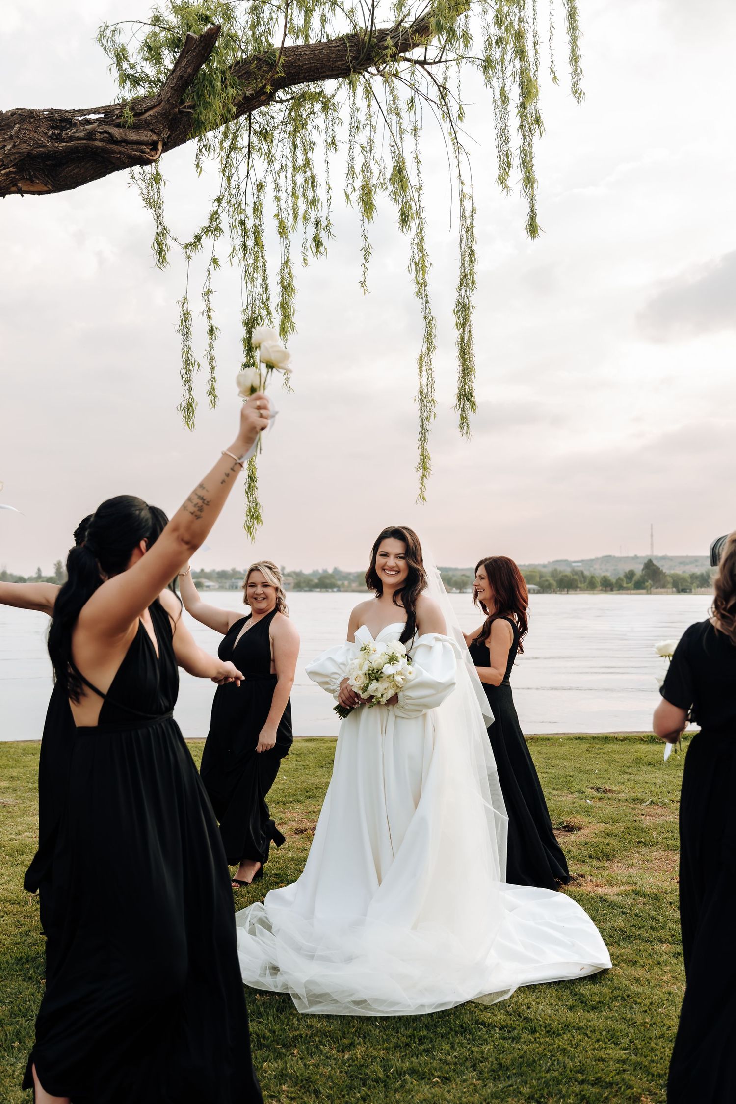 Bridesmaids in black dresses playfully lift bride in white gown under willow tree by lakeside during wedding photos.