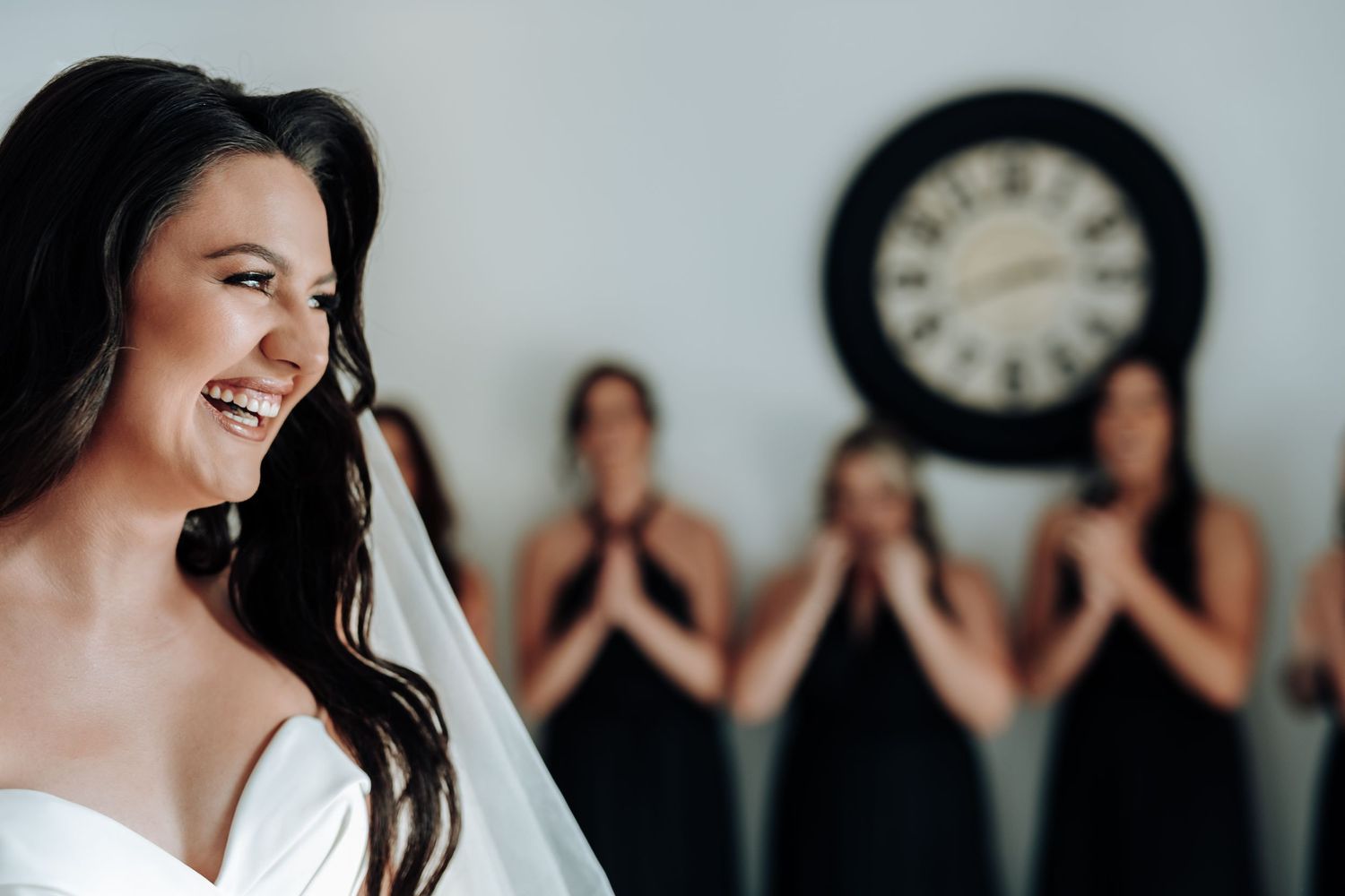 A joyful bride in white smiles while bridesmaids in black dresses react emotionally in the background with a clock on the wall.