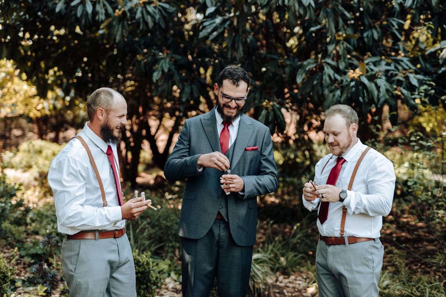 Groomsmen in light gray suits and dark red ties chat outdoors against a leafy background.
