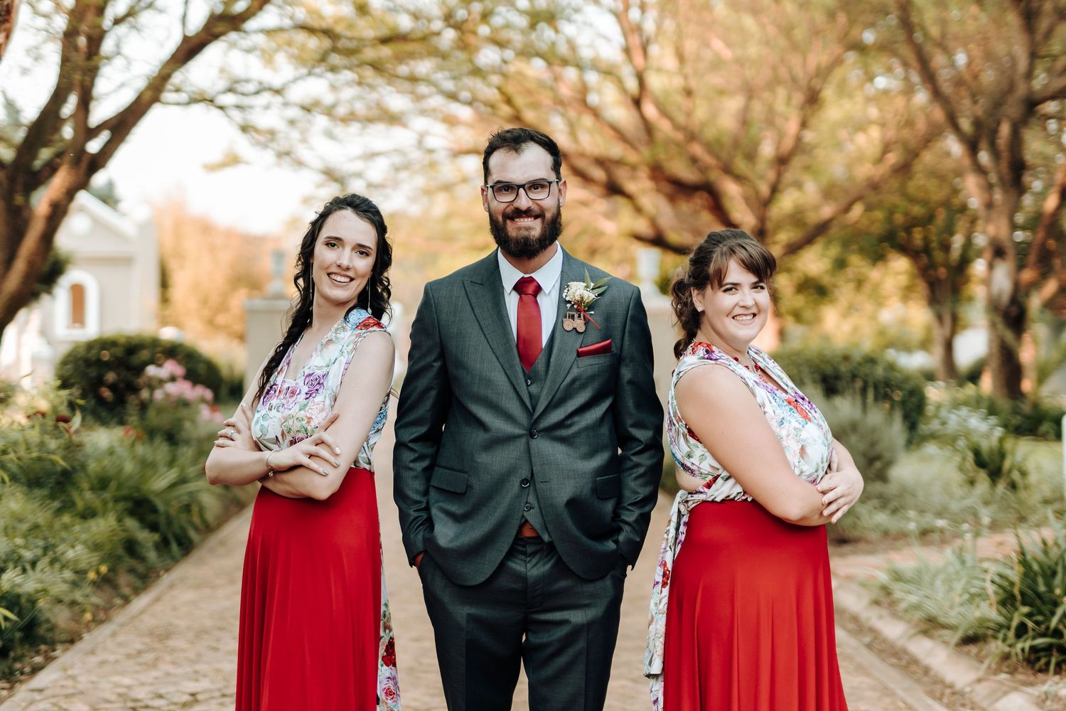 Wedding party poses playfully on brick path with two attendants in red dresses and dark grey suit in center.