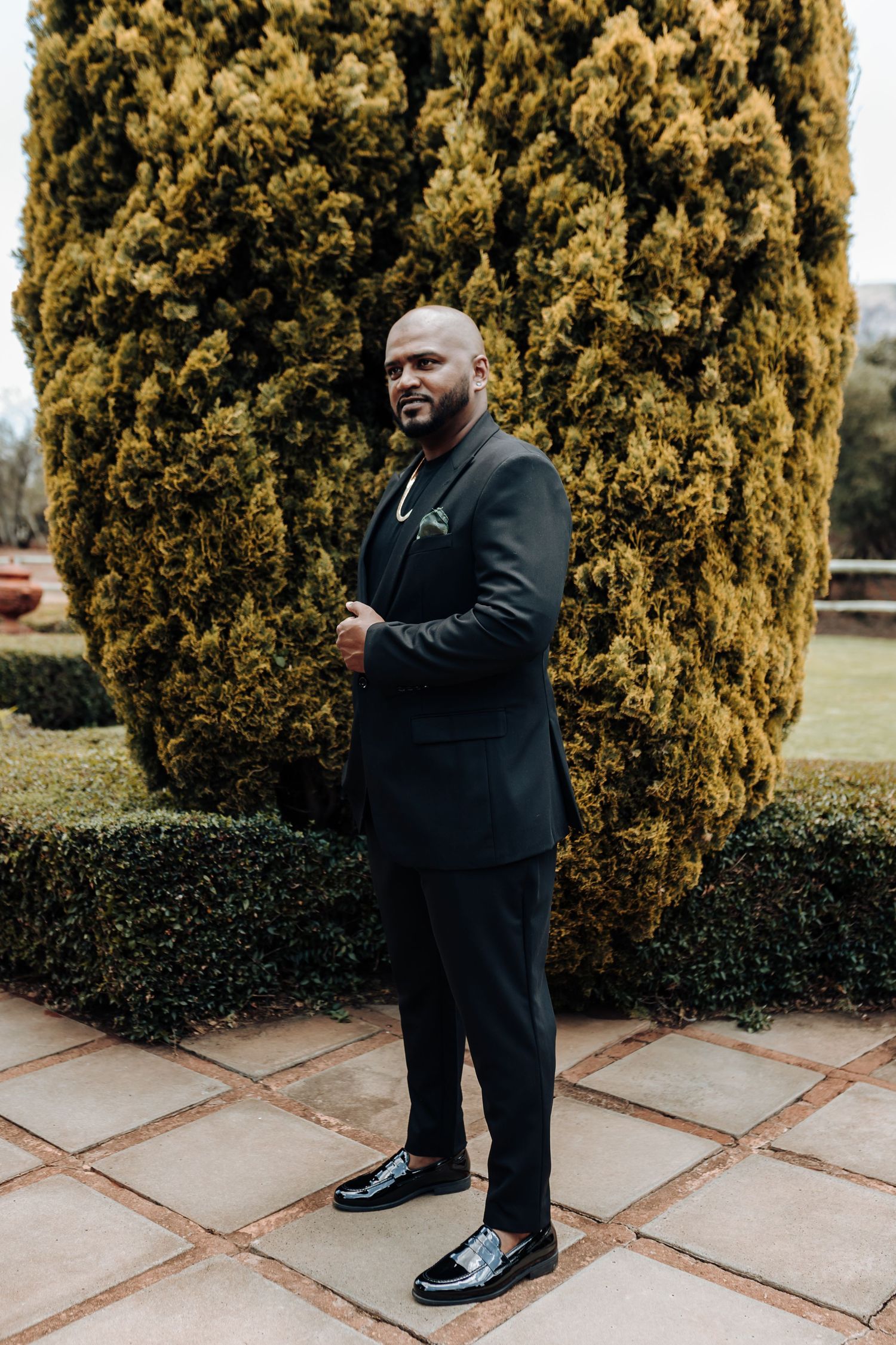 Well-dressed person in black suit standing against golden cypress tree and manicured hedges in formal garden setting.
