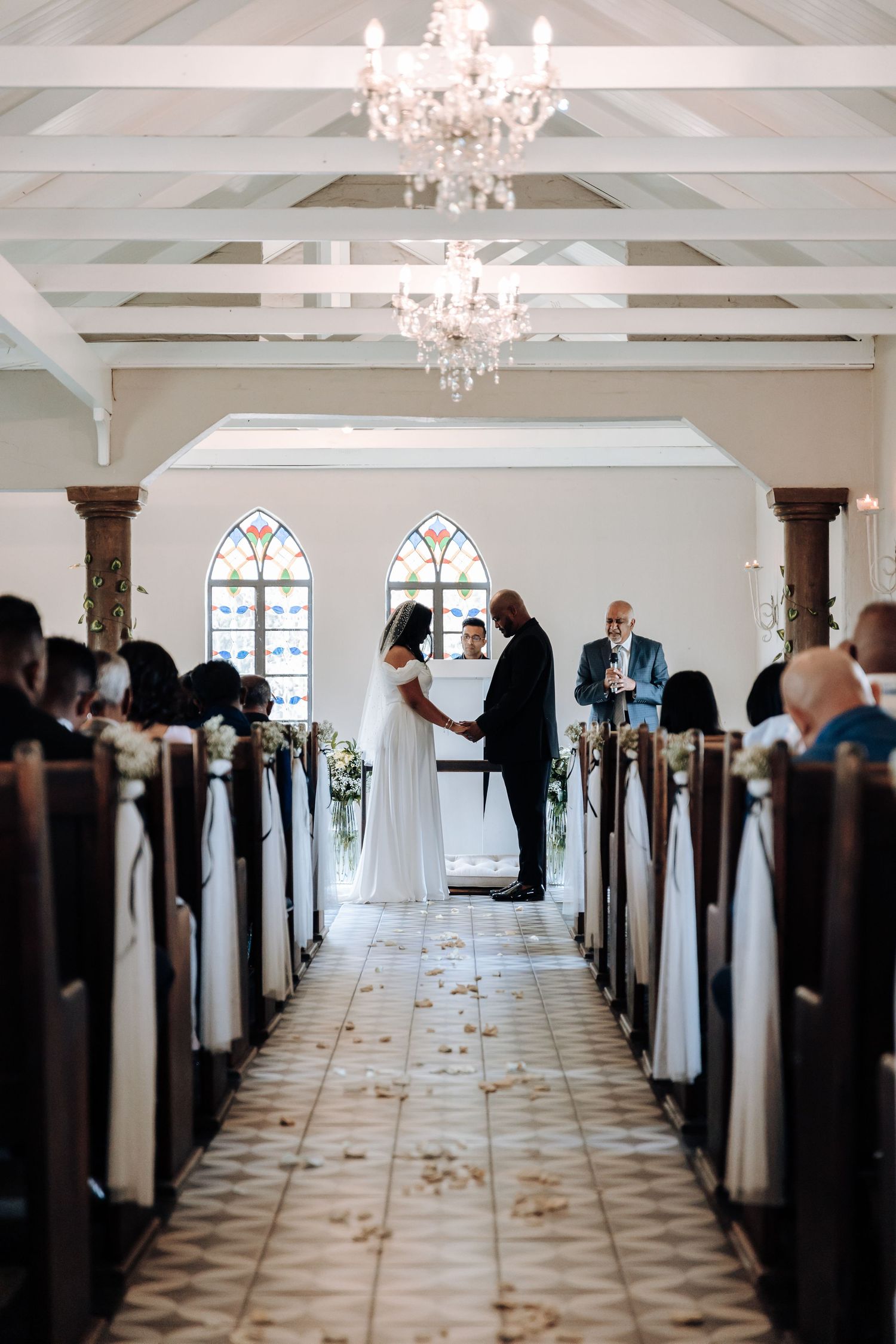 A wedding ceremony in a chapel with elegant chandeliers and white-decorated pews along the aisle.