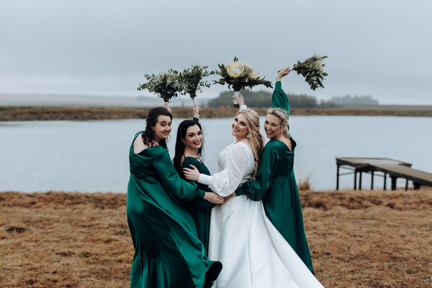 Wedding party in emerald dresses huddle together by waterfront on cloudy day.