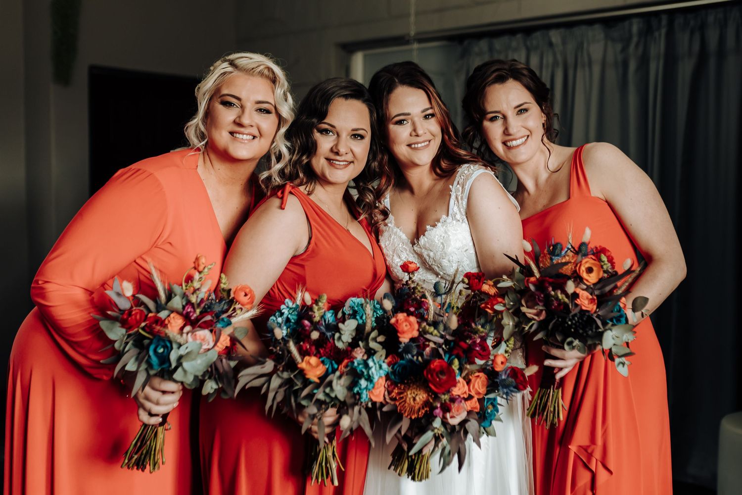 Wedding party smiling while holding colorful bouquets featuring coral and turquoise florals against grey backdrop.