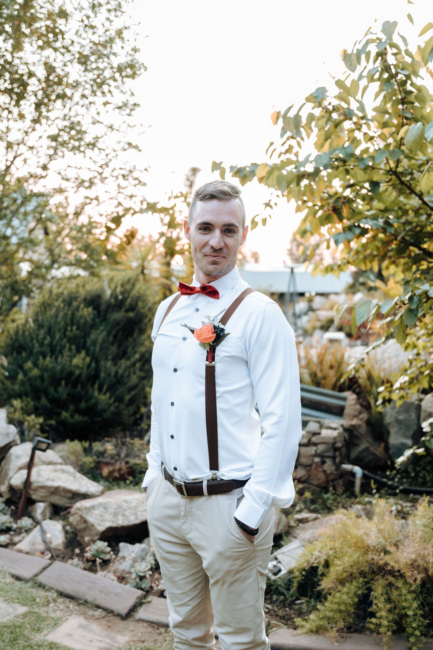 Groomsman in formal attire with red bow tie poses against garden backdrop.