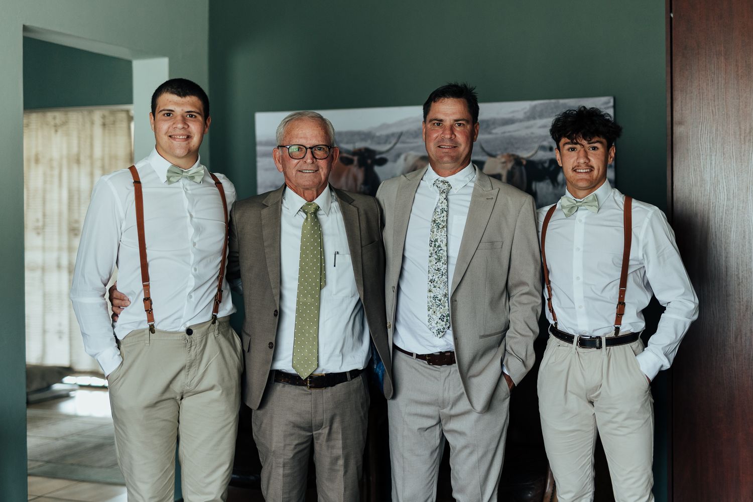 Men in coordinating light colored suits and suspenders pose against dark green wall.