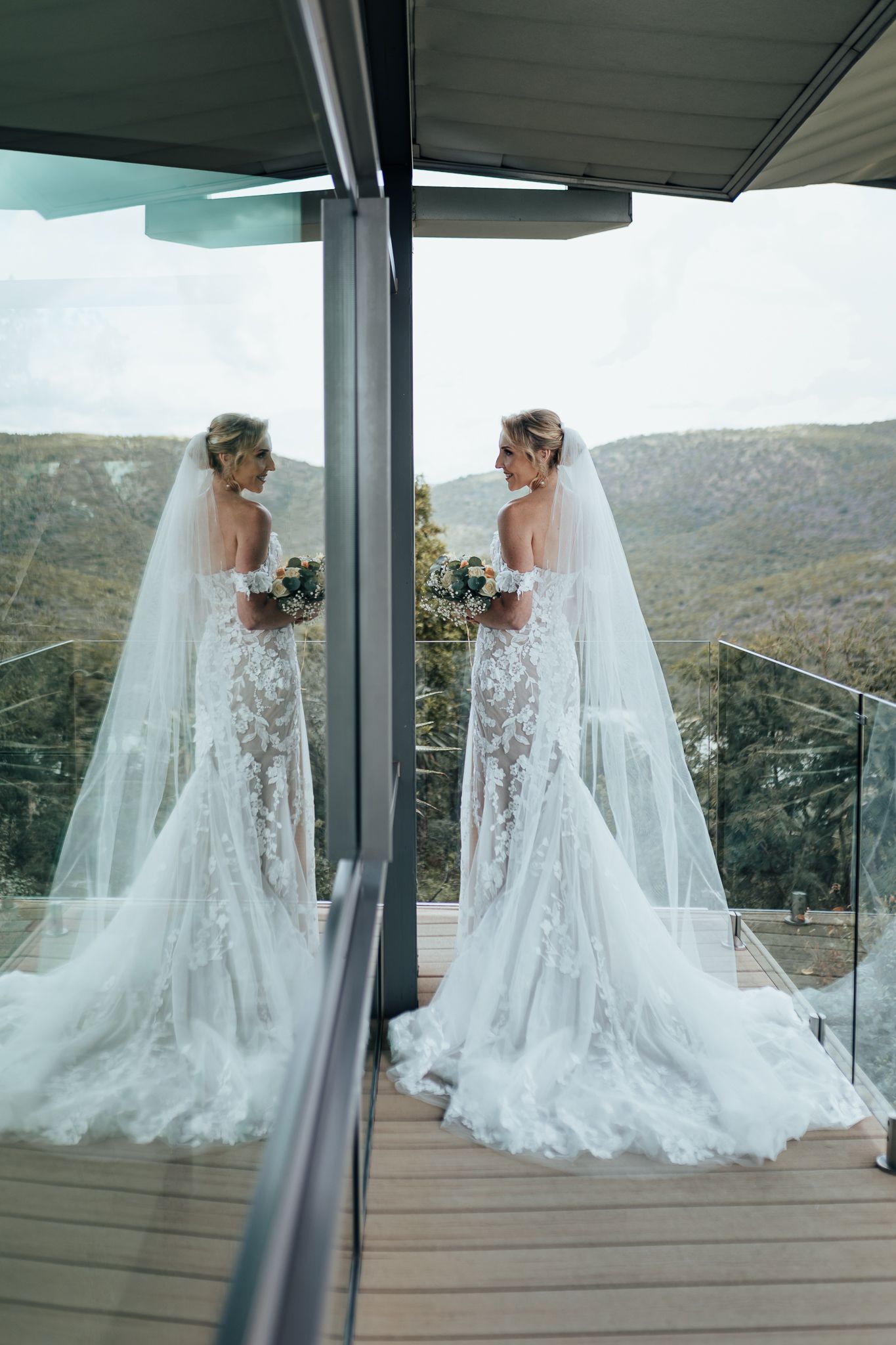 Reflection shot of bride in lace gown with long veil on modern deck with mountain views.