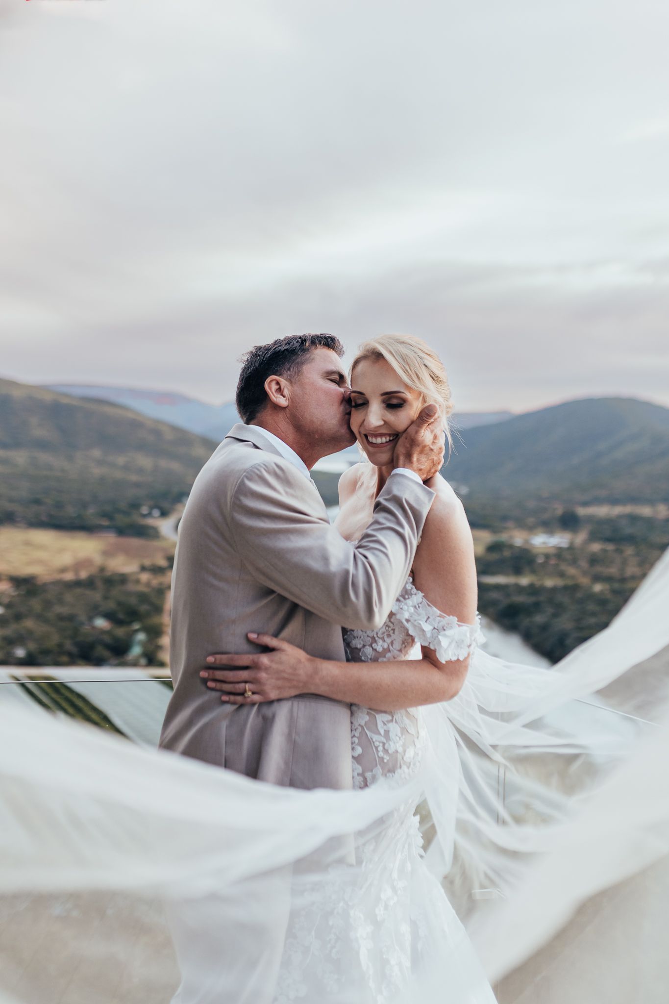 Romantic wedding moment captured with flowing veil against mountain landscape.