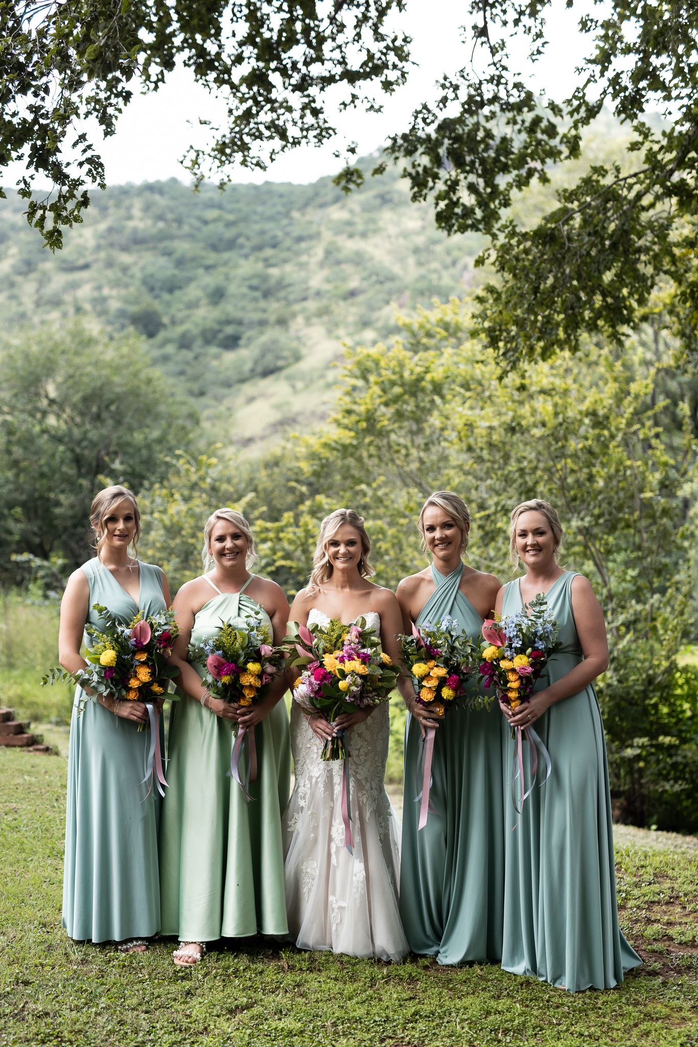 Wedding party in mint green dresses poses with colorful bouquets against a mountain backdrop.