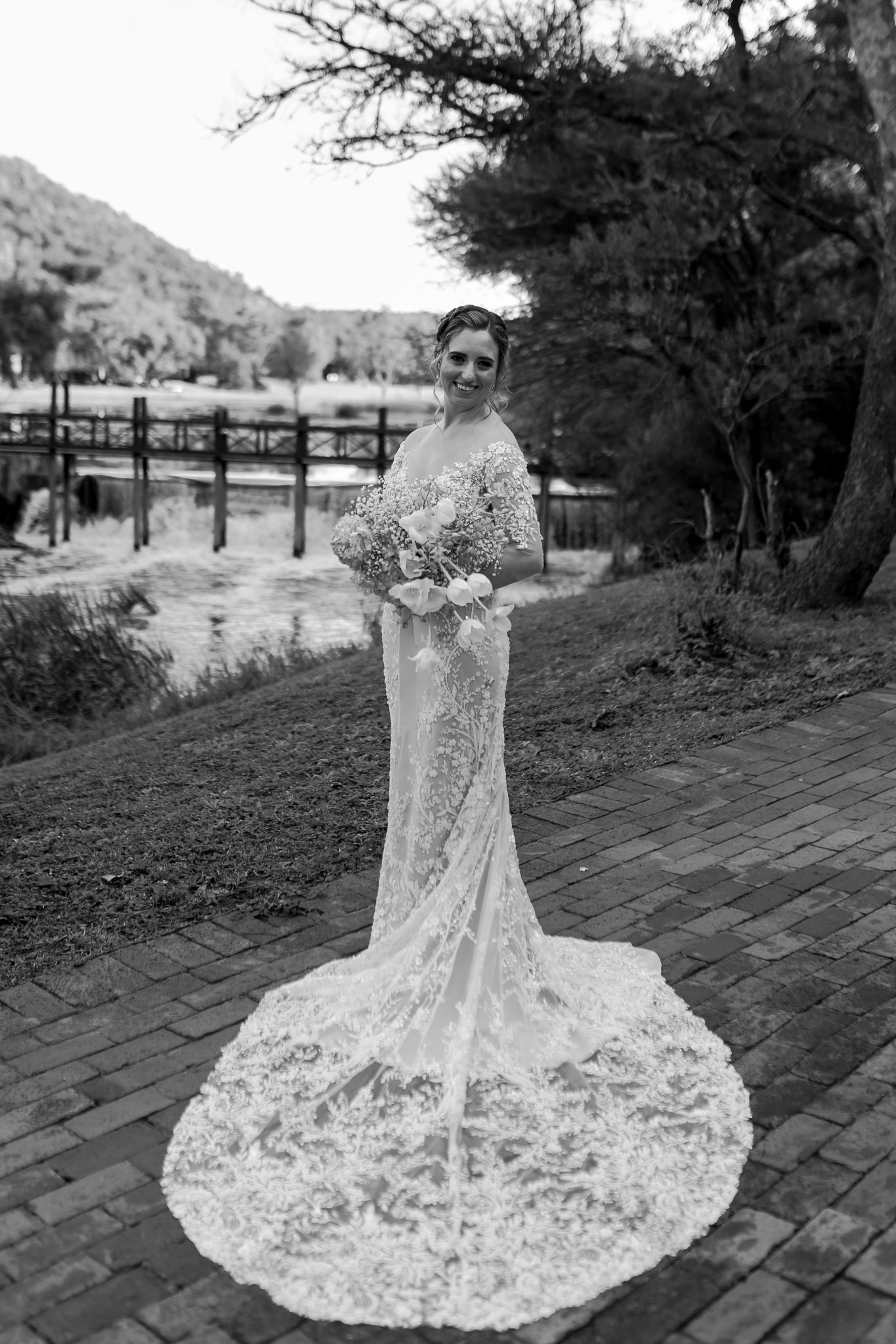 Black and white portrait showcasing intricate lace wedding gown with cathedral train on brick pathway.