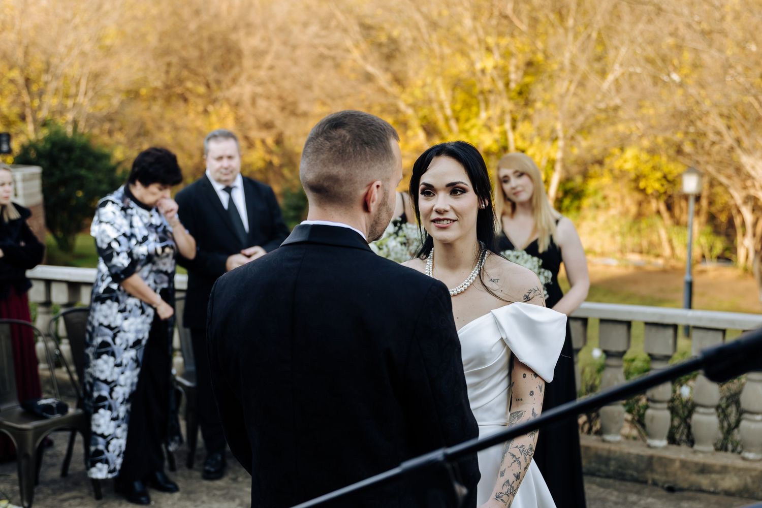 A couple exchanges vows during an autumn outdoor wedding ceremony with guests watching in the background.