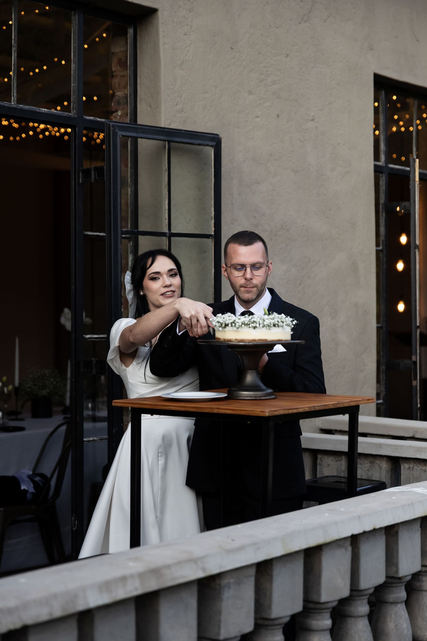 A couple in formal wedding attire stand together at an elegant venue with twinkling lights in the background.