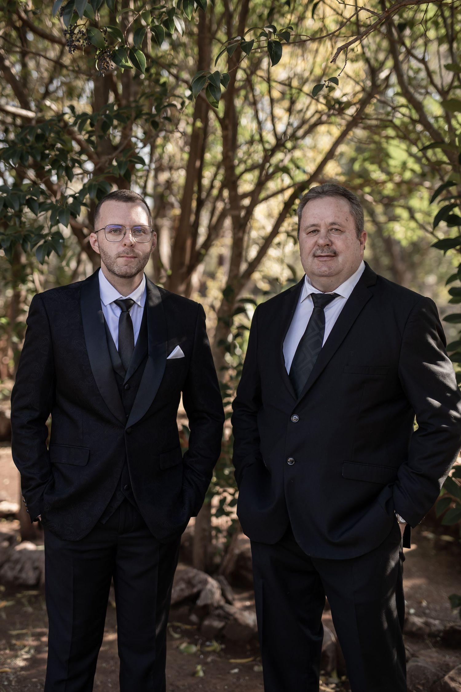 Two men in black suits and ties stand together outdoors in a wooded garden setting.