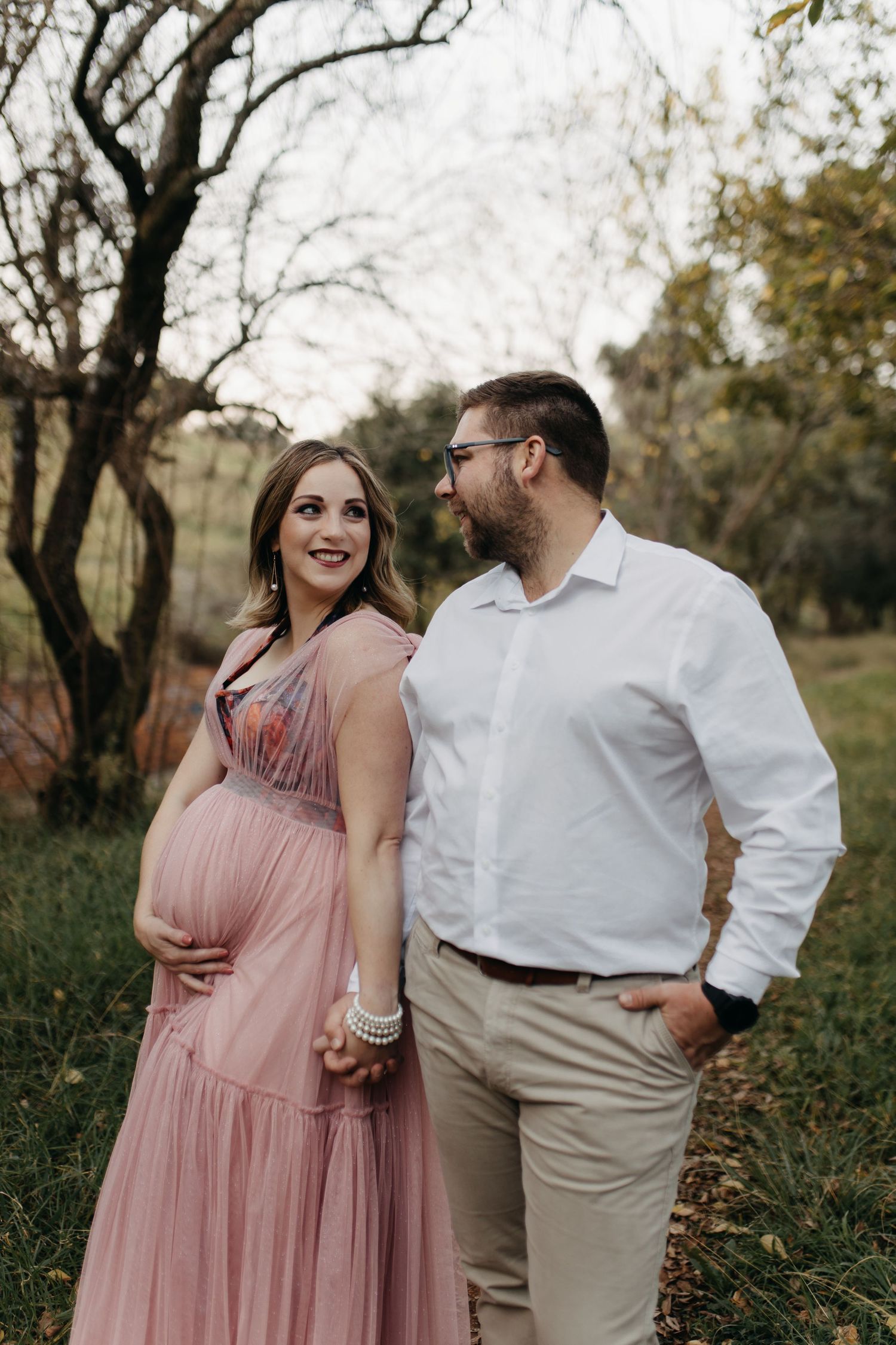 A couple shares tender moments in a natural outdoor setting, with her in a pink maxi dress and him in casual attire.