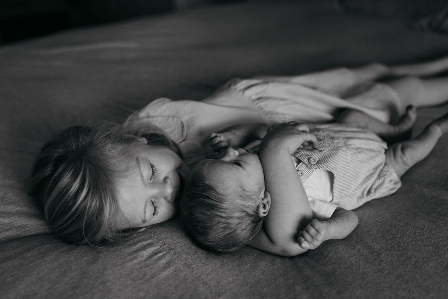 Two sleeping children cuddle close together on soft bedding in a tender black and white photograph.