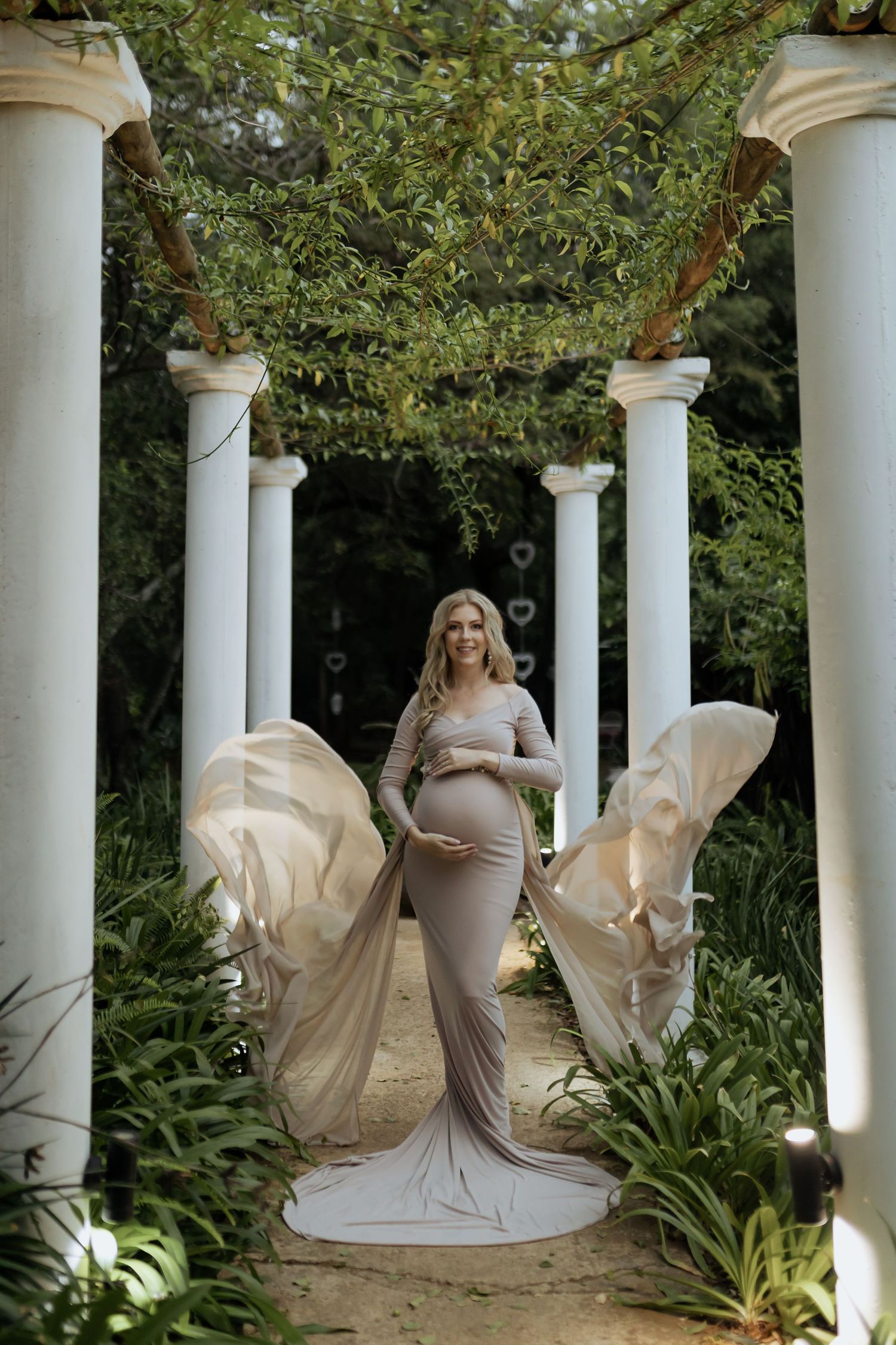 Maternity photo shoot in an elegant garden setting with white columns, featuring flowing beige dresses in multiple poses.
