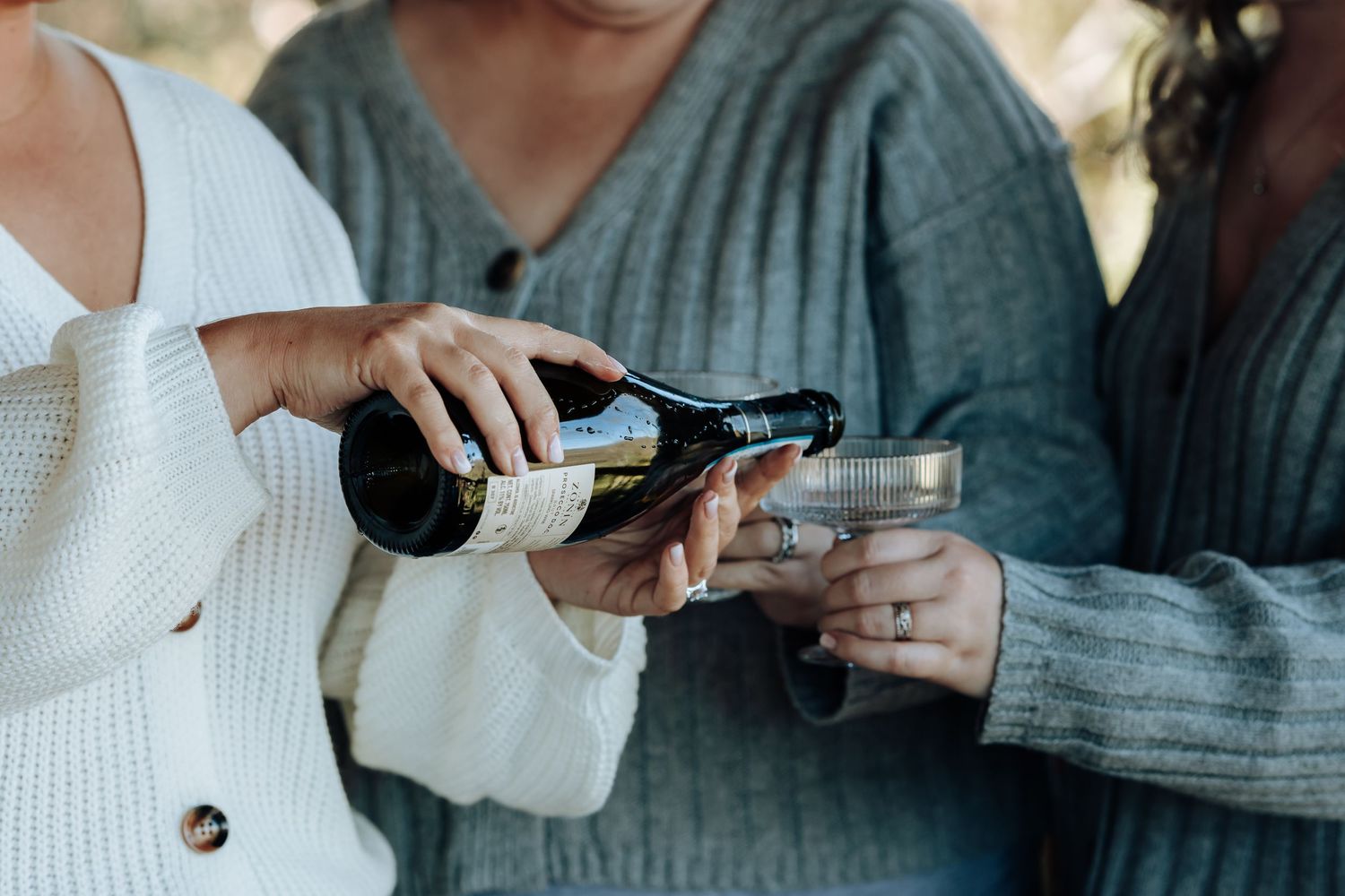 Two women in cozy sweaters pouring sparkling wine into glasses outdoors at a casual celebration.