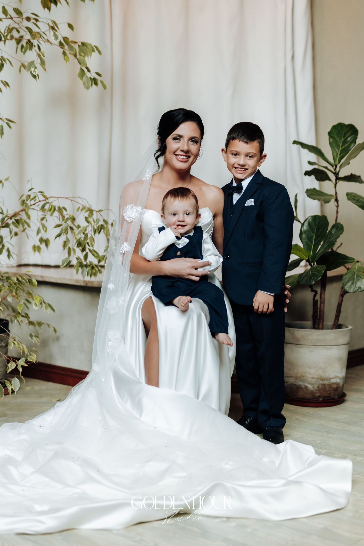 Bride in white gown holds infant while posing with young boy in navy suit at a wedding celebration.