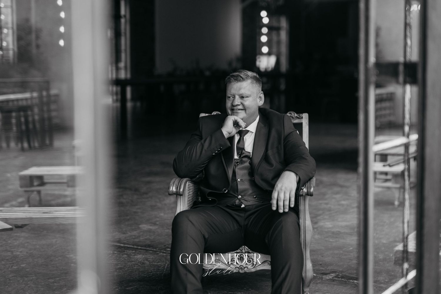 Black and white photo of a smiling man in a suit sitting in a chair at an elegant wedding venue.