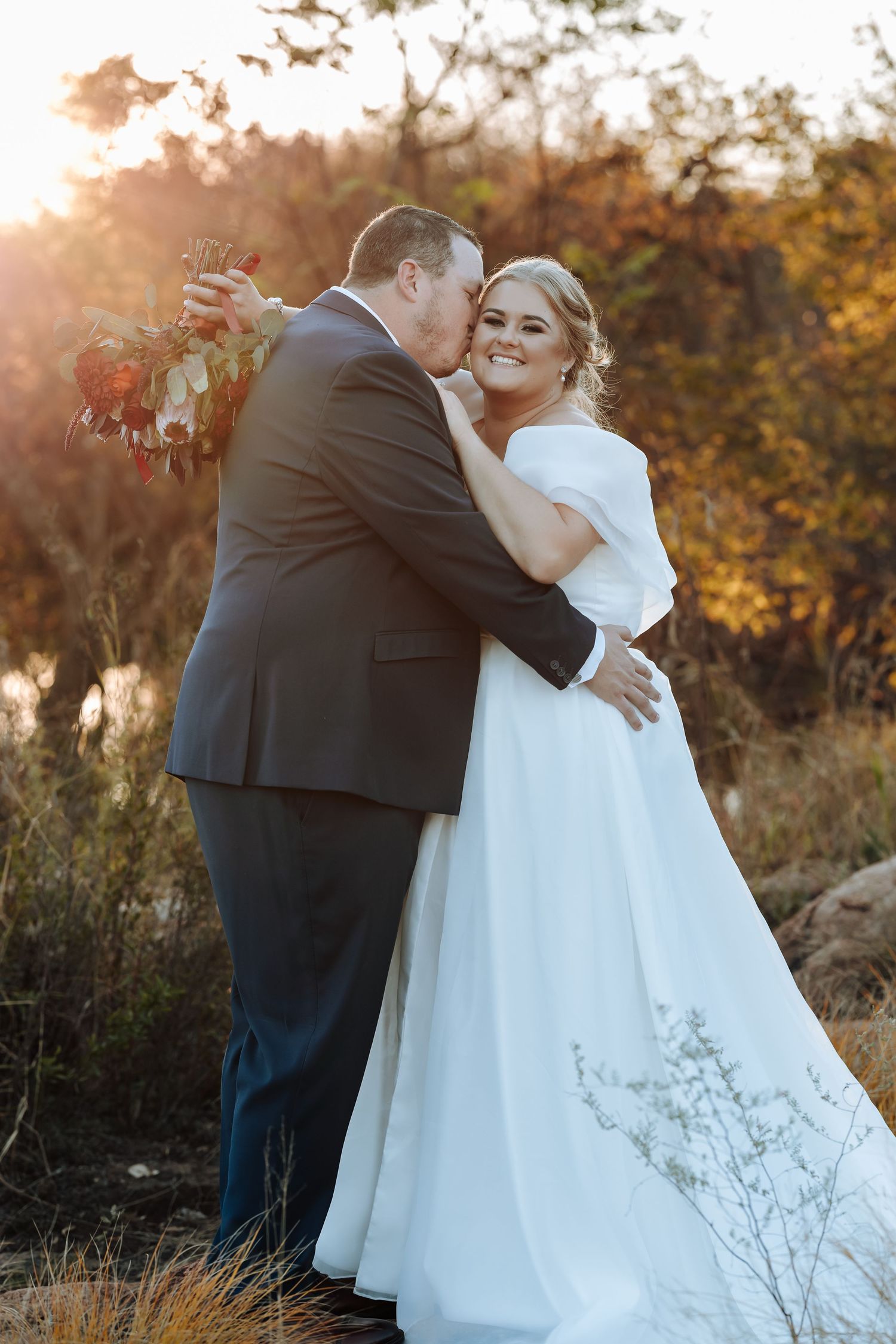 A bride in a white gown and groom in a dark suit share a romantic kiss during a golden sunset outdoor wedding.