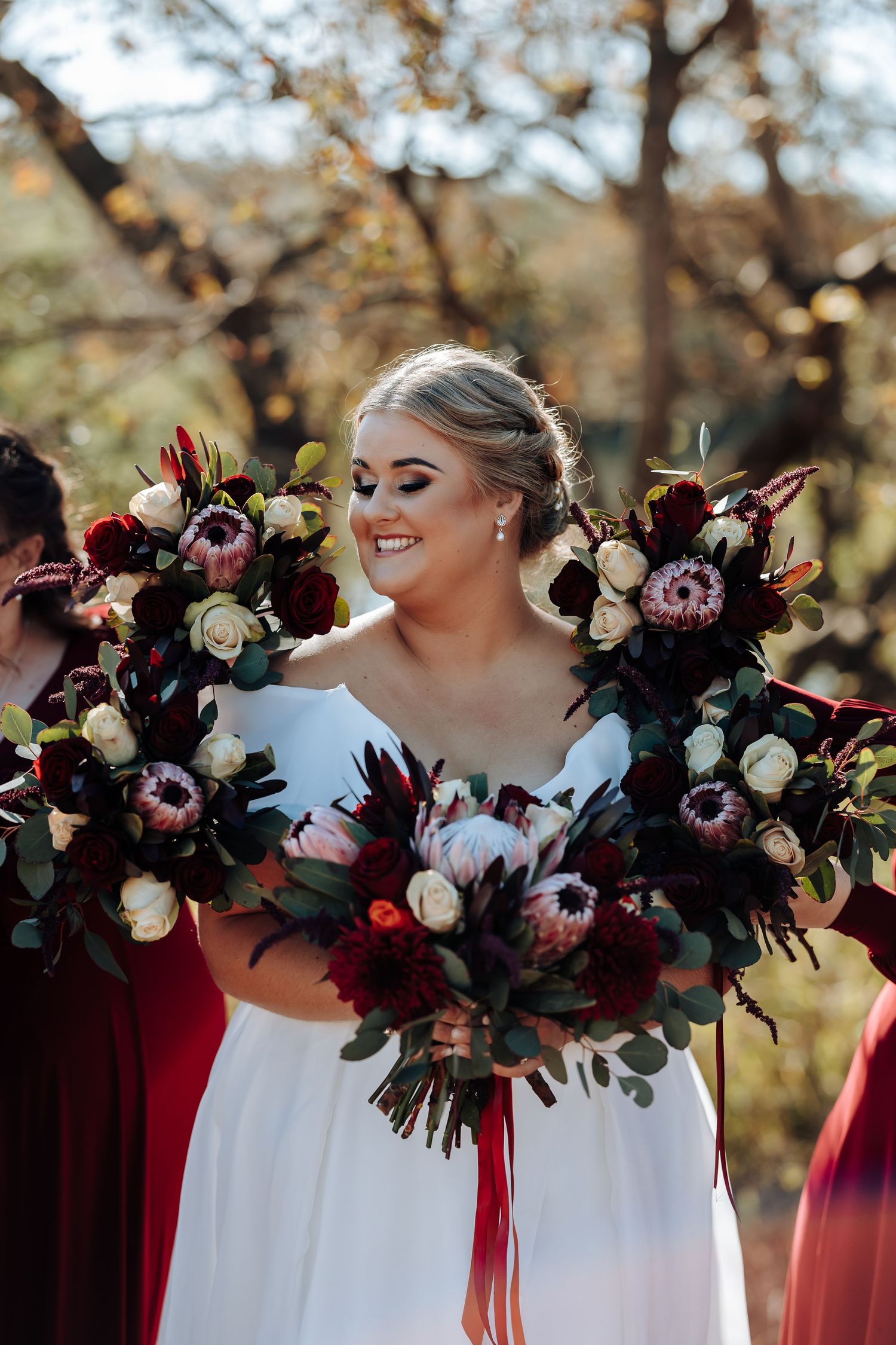Smiling bride in white gown holding large burgundy and cream floral bouquets with proteas outdoors.