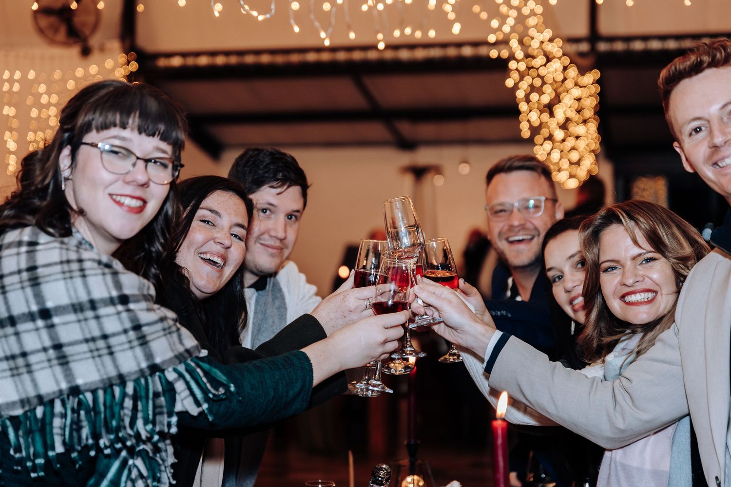 Group of friends toasting with wine glasses at a festive dinner party with warm string lights in background.