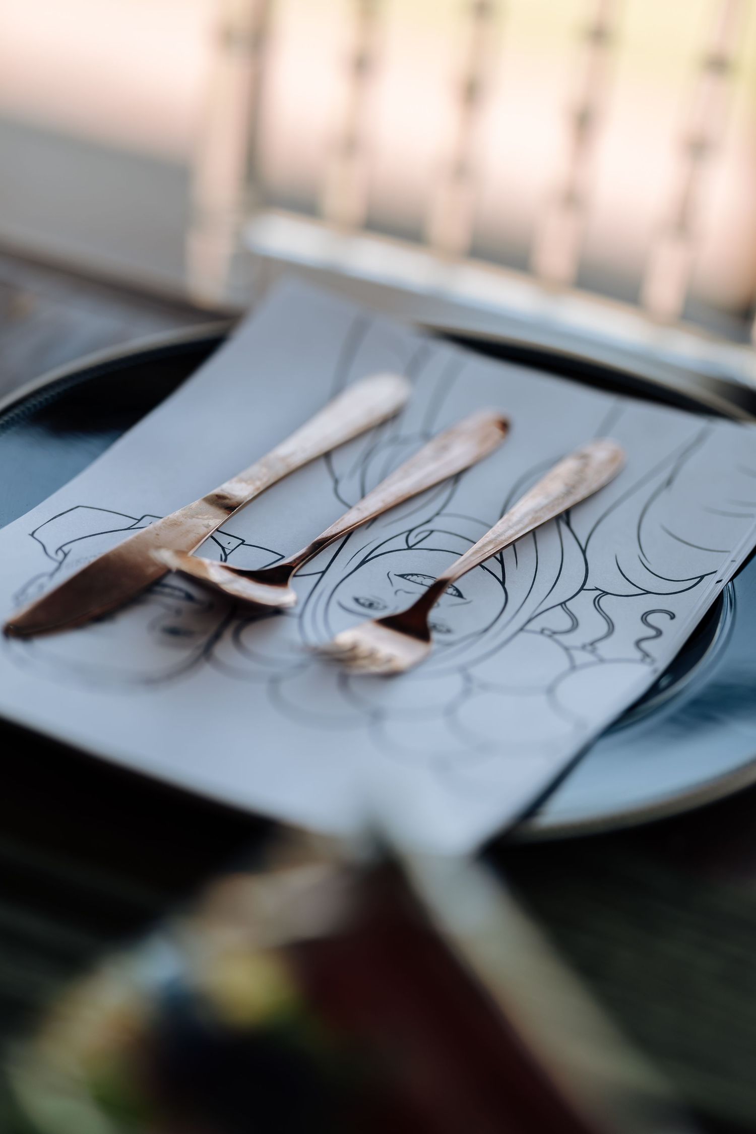 Rose gold cutlery set with knife and forks resting on decorative paper plate with artistic line drawing.