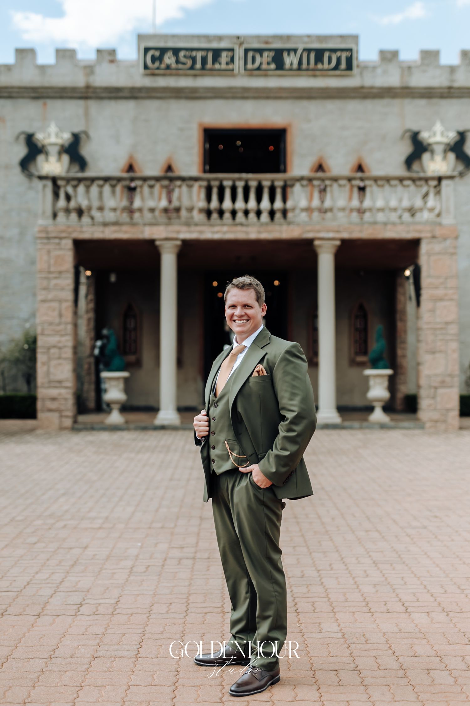 Groom in olive green suit poses confidently outside Castle De Wildt on wedding day.