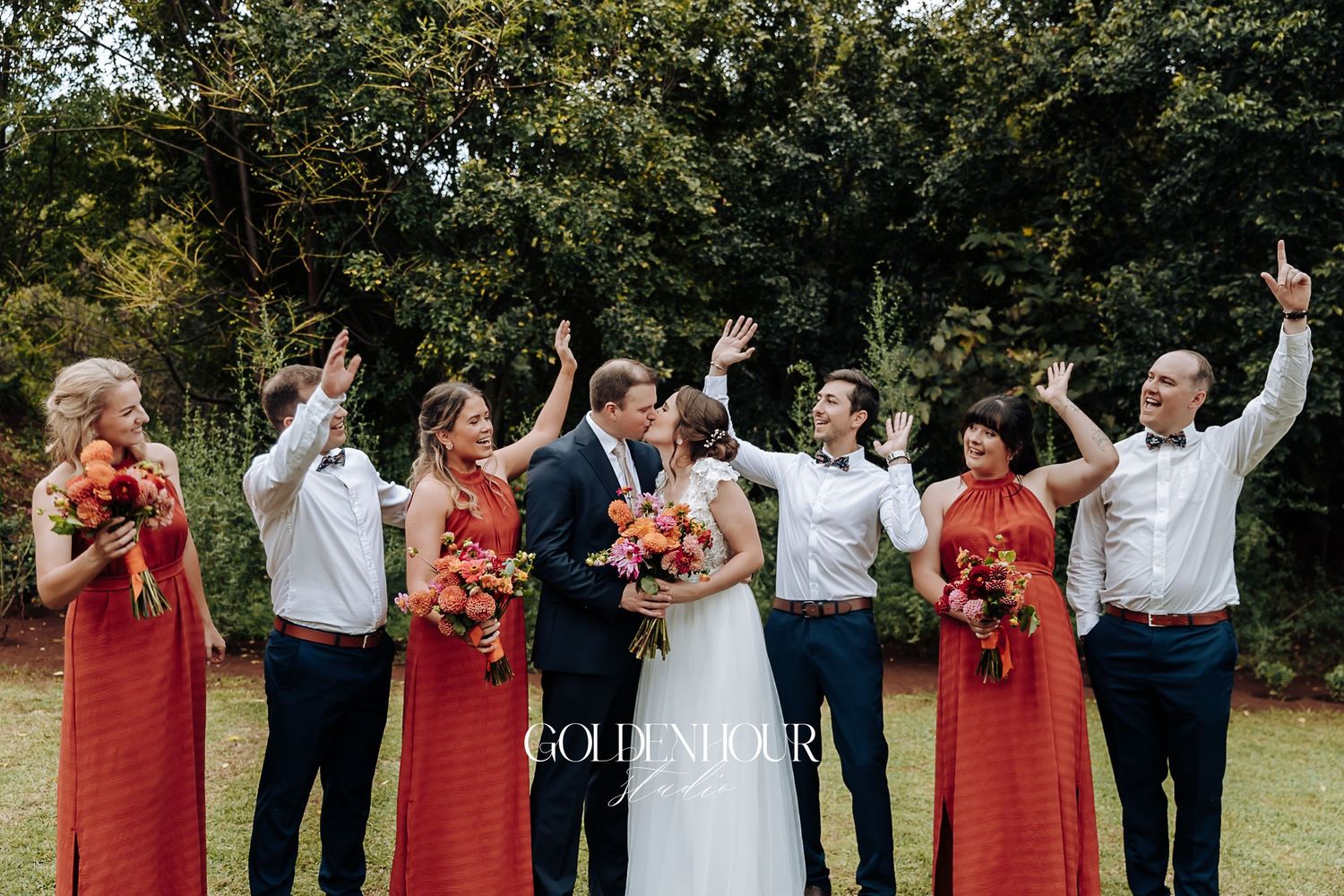 Wedding party in red dresses and white shirts celebrating with raised arms behind kissing couple.