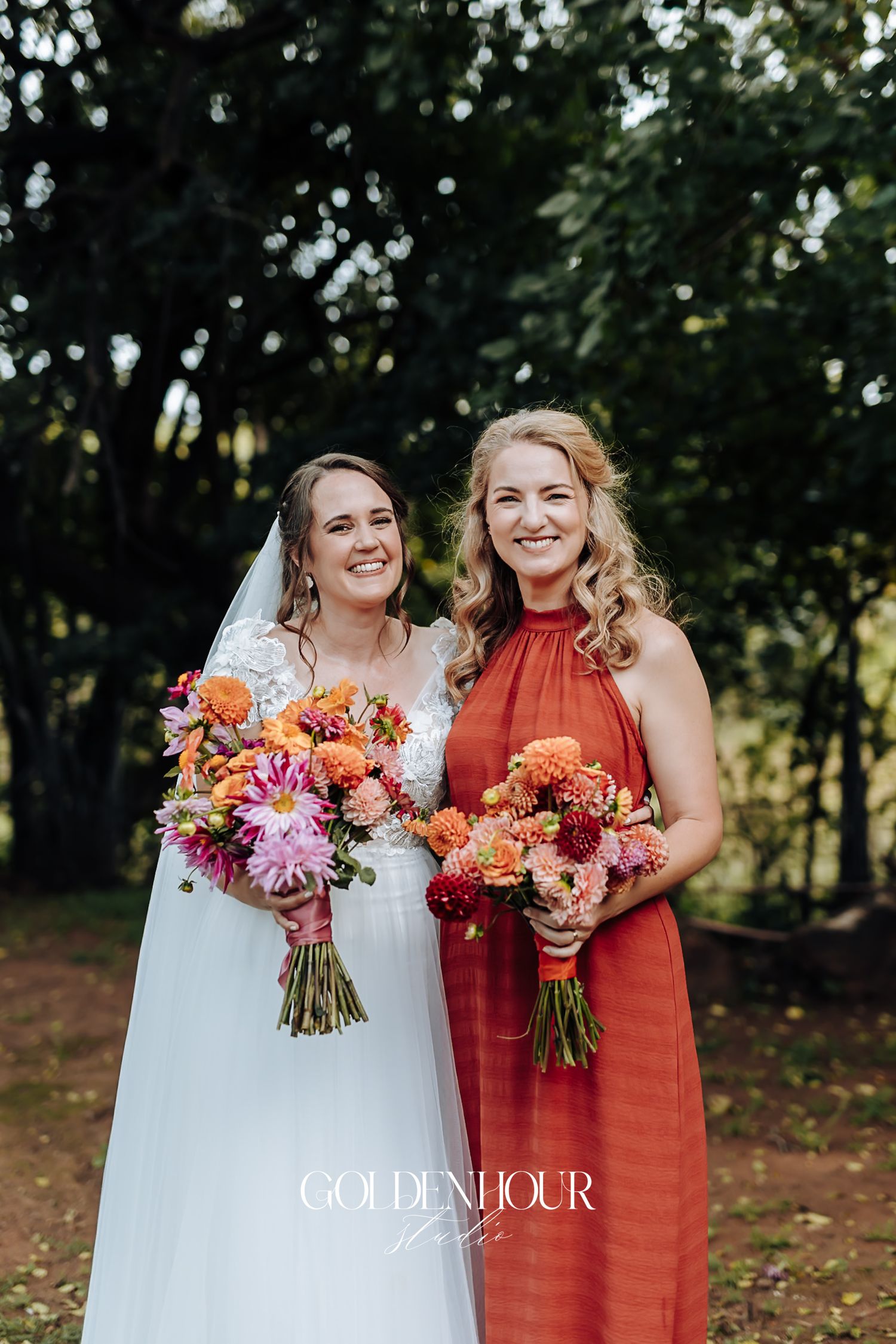 Bride in lace wedding dress and bridesmaid in red gown holding colorful bouquets outdoors.