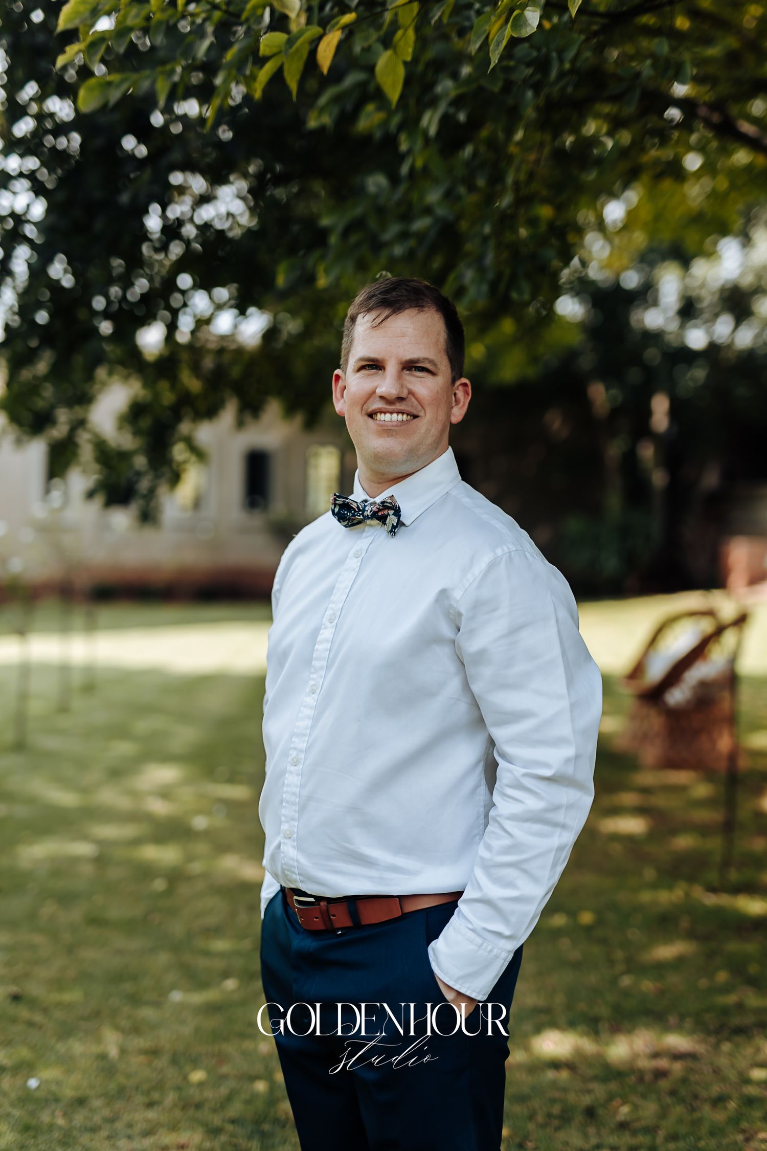 Smiling groom in white shirt and floral bow tie posing outdoors at wedding venue.
