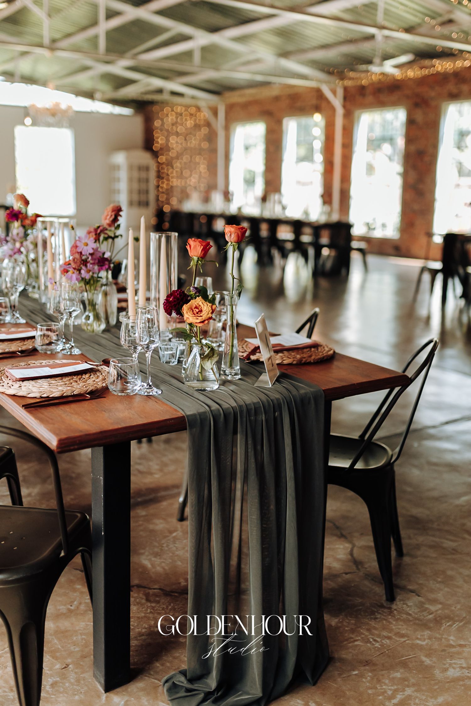 Elegant wedding reception table with gray runner, colorful floral centerpieces, candles, and rustic wood tables in a brick venue.