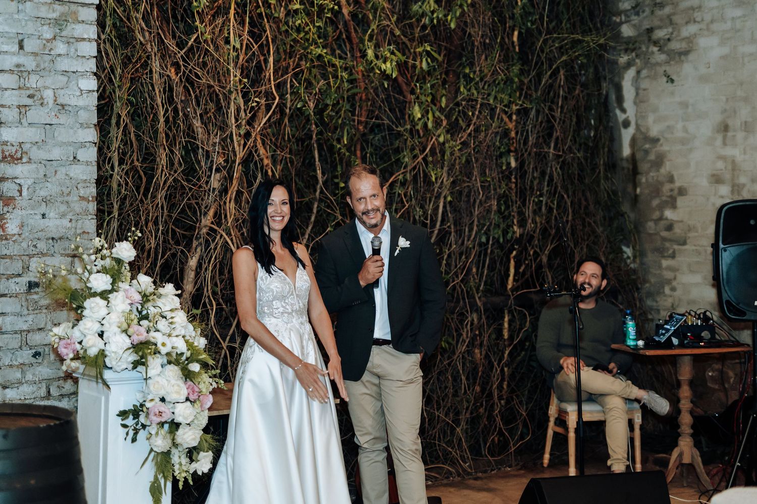 Bride and groom smiling together at wedding reception with floral arrangements and rustic vine backdrop.